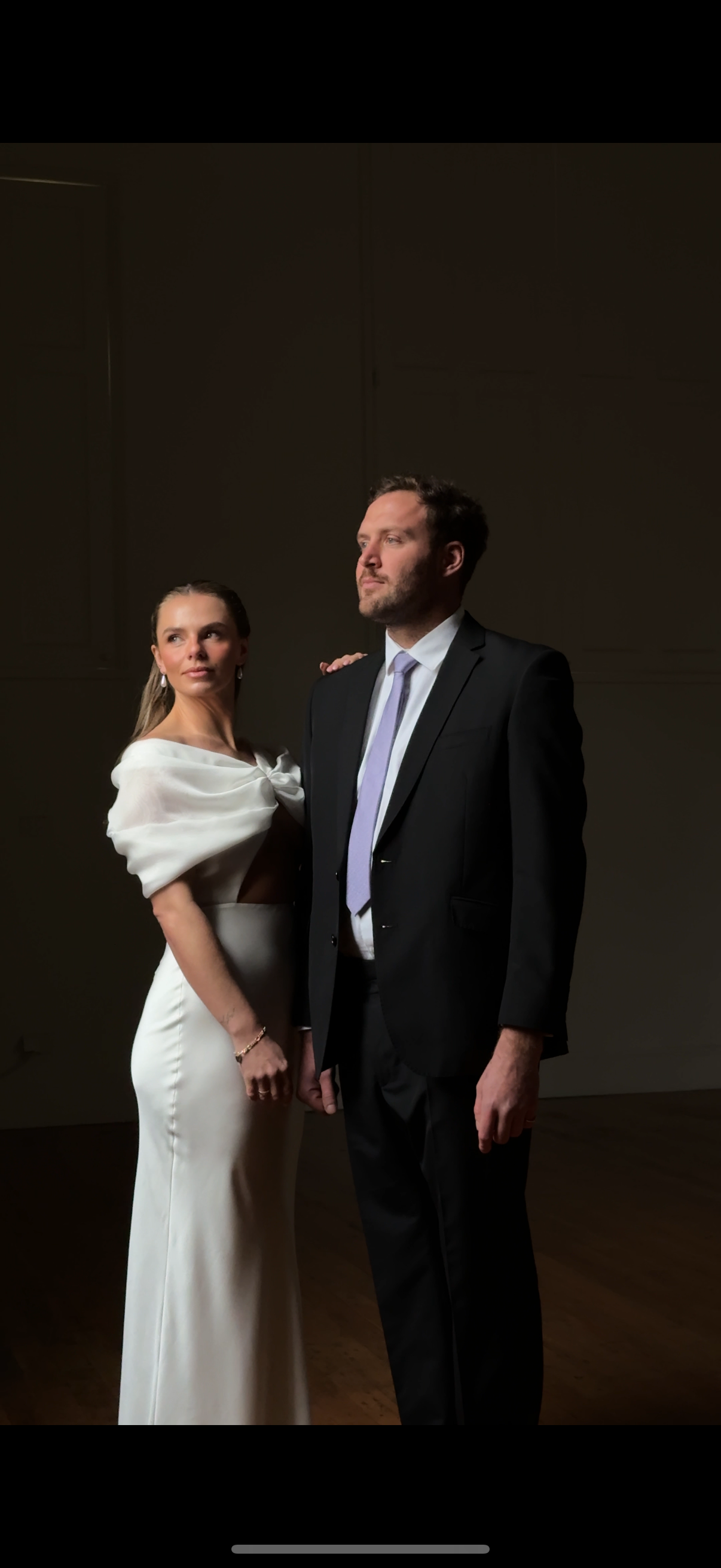 A newly married couple stands together in a dark room, spotlight illuminating them as they look out a window, faces turned away from the camera 