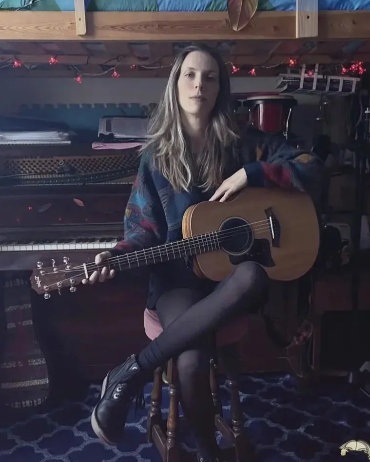 A young woman with long wavy hair sitting on a wooden chair and holding an acoustic guitar, in a room with a piano and string lights.