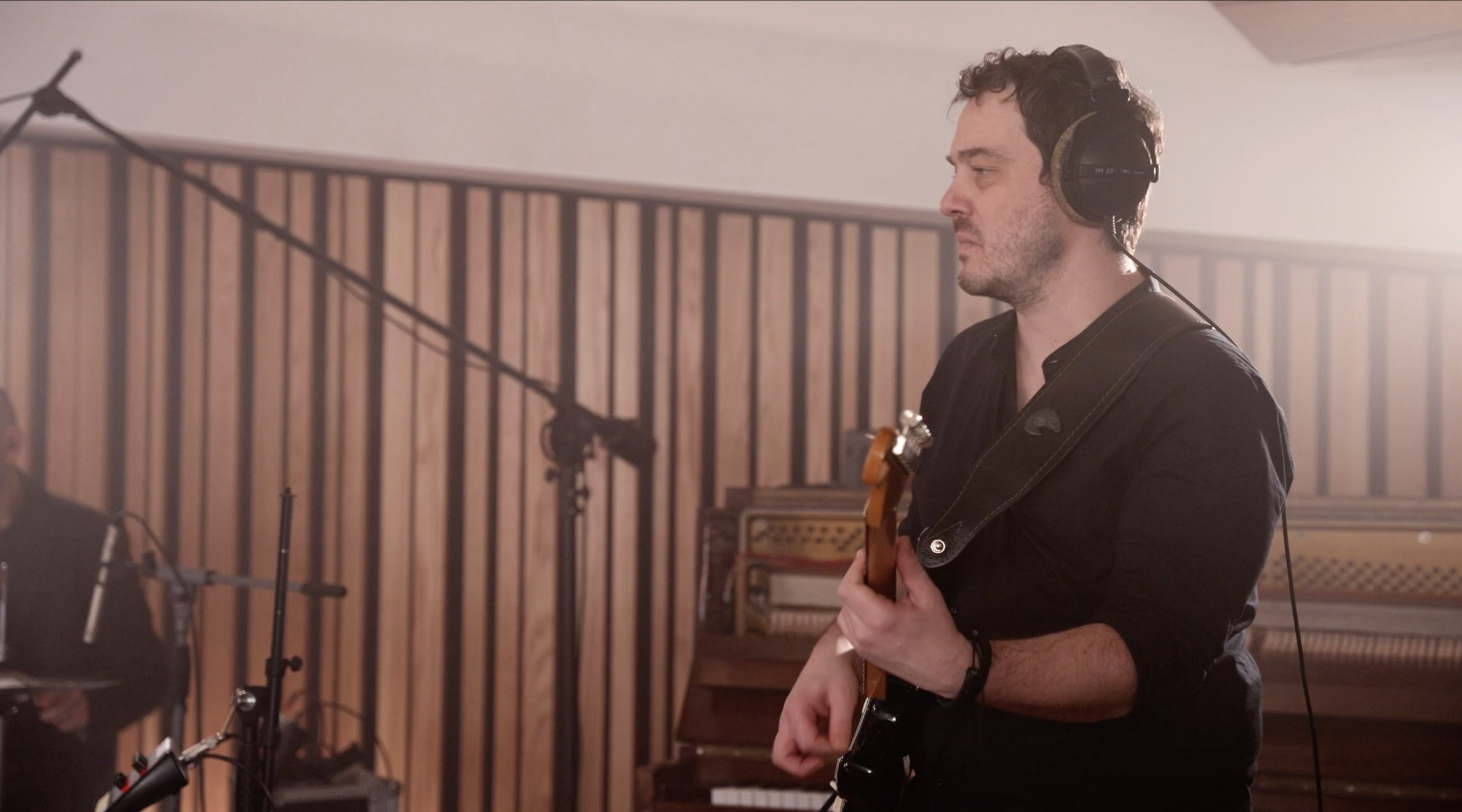 A man wearing headphones playing an electric guitar in a recording studio with wooden panel walls and a piano in the background.