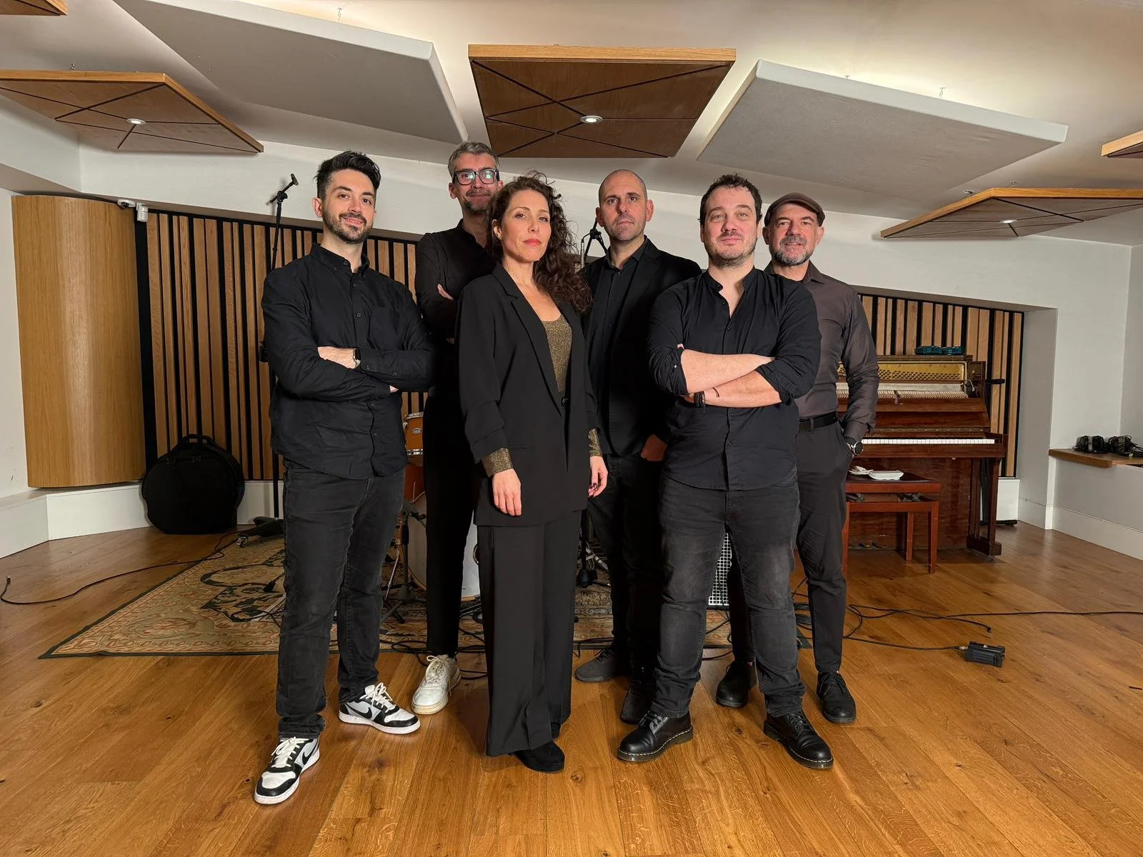 Group of six people, five men and one woman, standing in a music studio with a piano and musical equipment in the background.