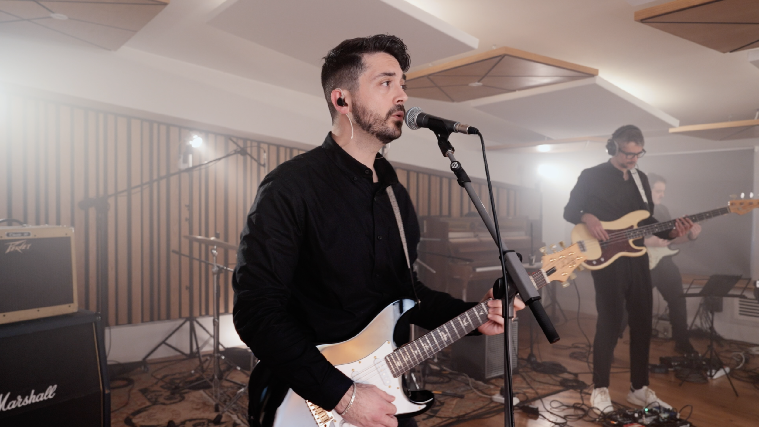 A band is performing in a studio, with a man singing into a microphone and playing an electric guitar, and other musicians in the background playing bass and guitar.