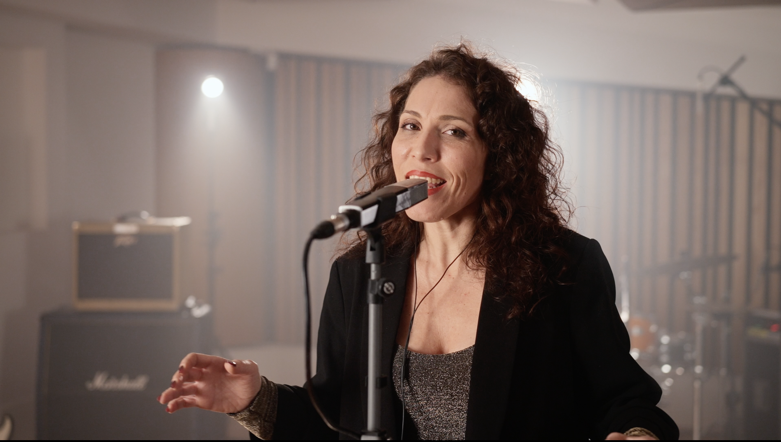 A woman with curly brown hair singing into a microphone in a music studio with amps and drums in the background.