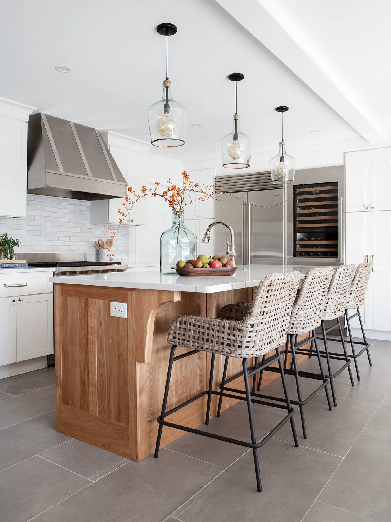 Modern kitchen with white cabinets, a wooden island with a white countertop, four wicker bar stools, pendant lights, a large glass vase with orange berries, and a tray of apples.