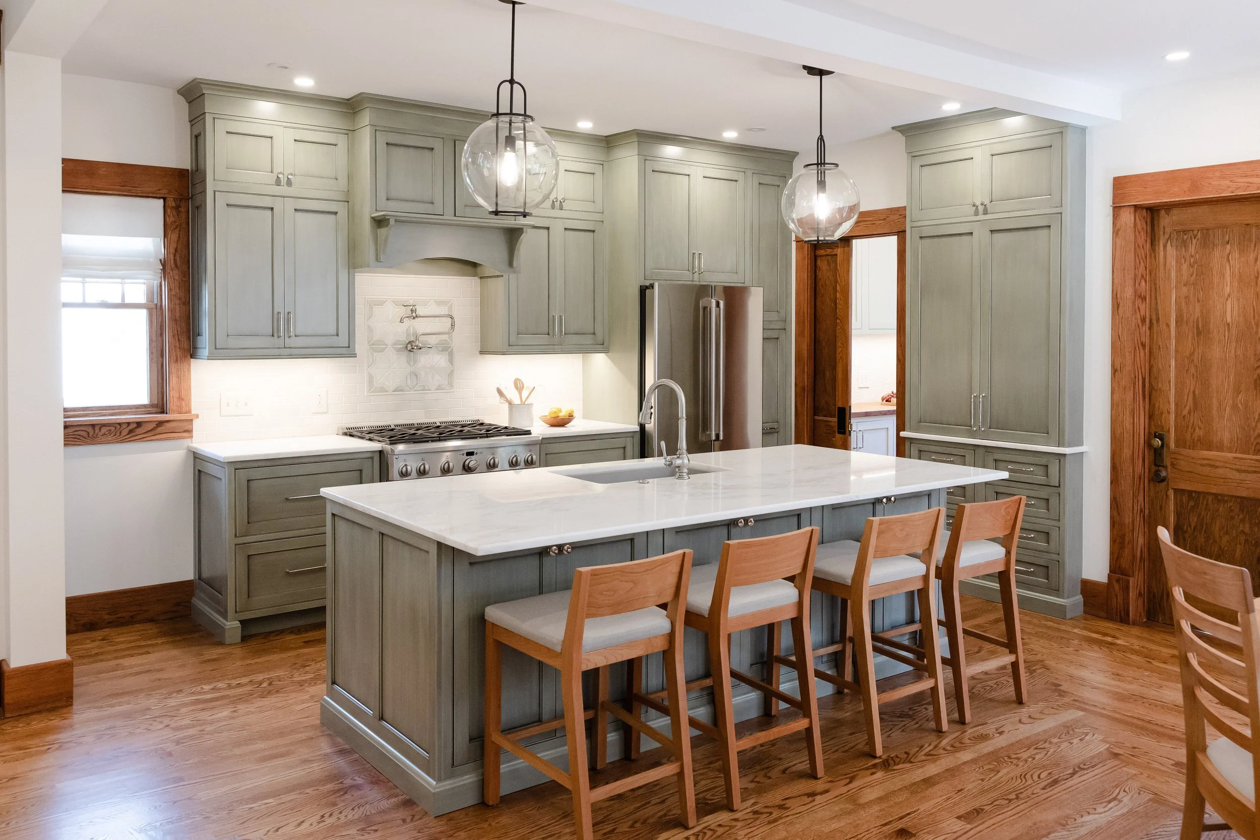 Modern kitchen with light green cabinets, white marble island, wooden chairs, stainless steel appliances, hardwood floors, and pendant lighting.
