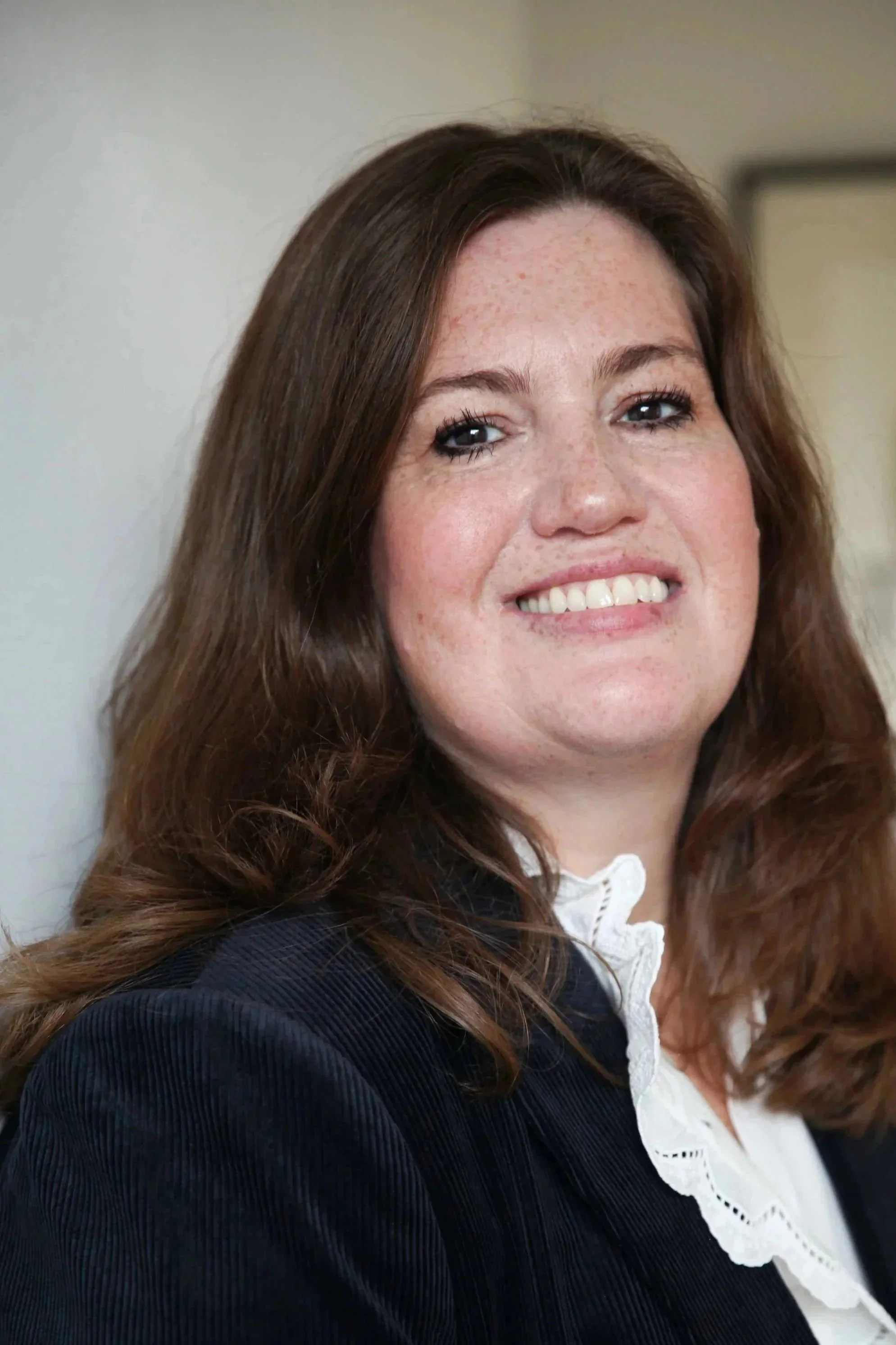 A woman with long, wavy brown hair smiling, wearing a black jacket and a white ruffled shirt, in a well-lit indoor setting.