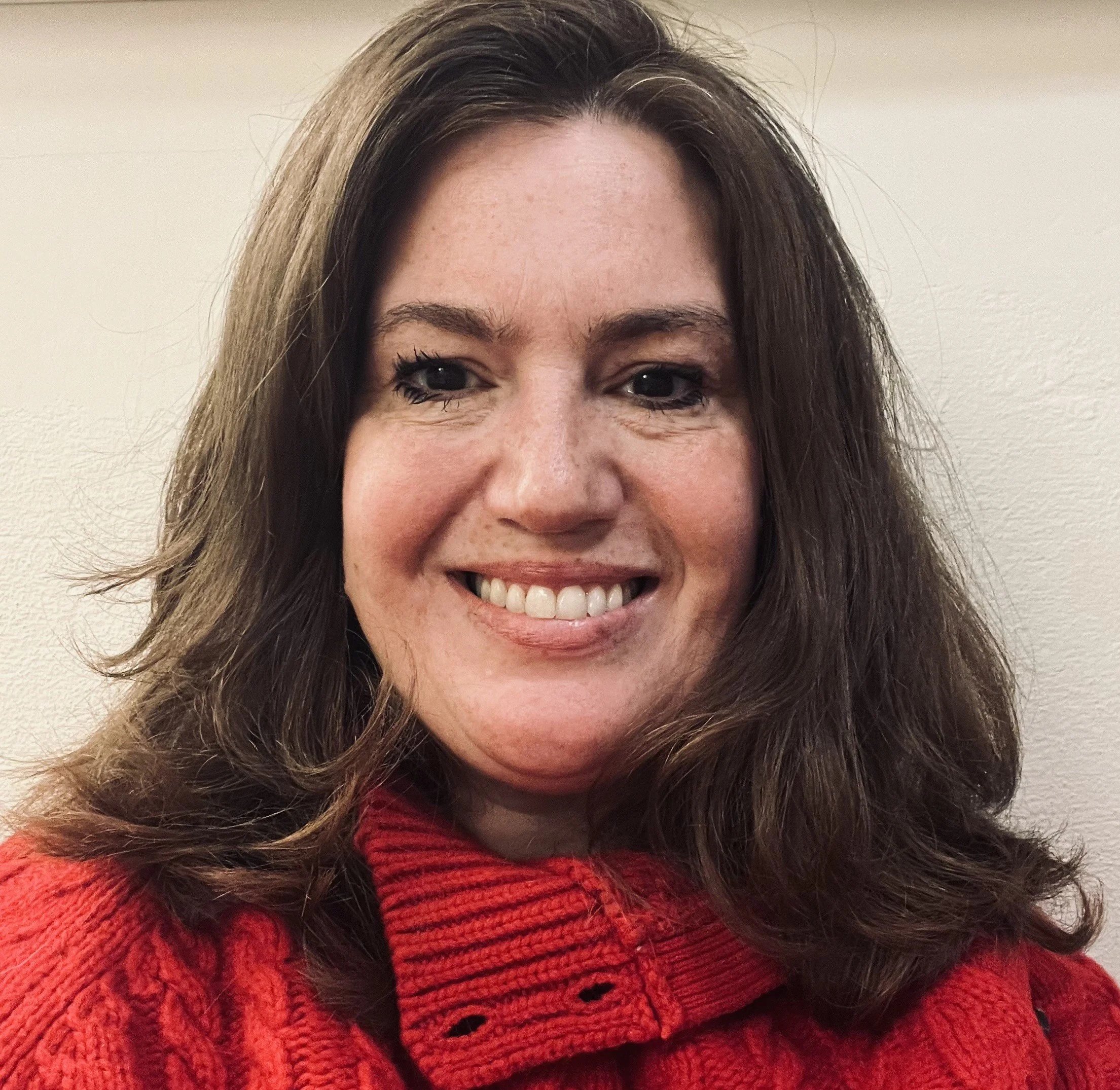 Close-up of a woman with shoulder-length brown hair, smiling, wearing a red sweater, standing against a plain beige wall.