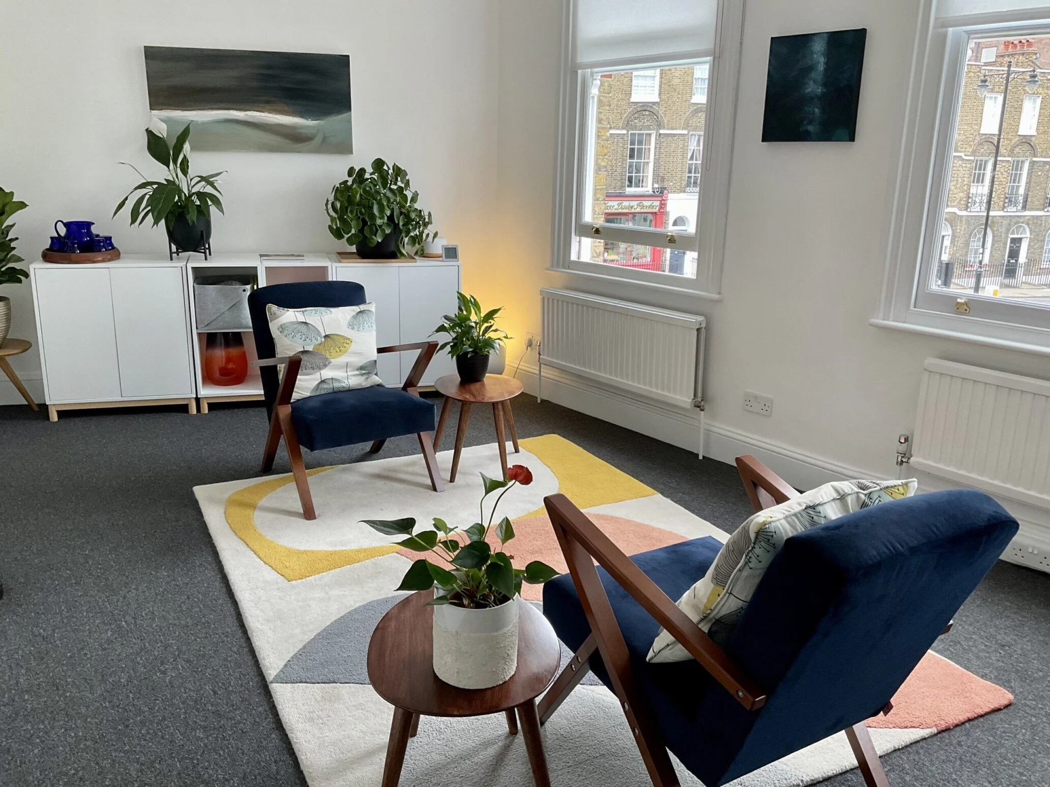A modern living room with two blue armchairs, a white rug with yellow, pink, and blue accents, a round wooden side table with a plant, a white storage unit with houseplants and decorative items, and large windows overlooking a cityscape.