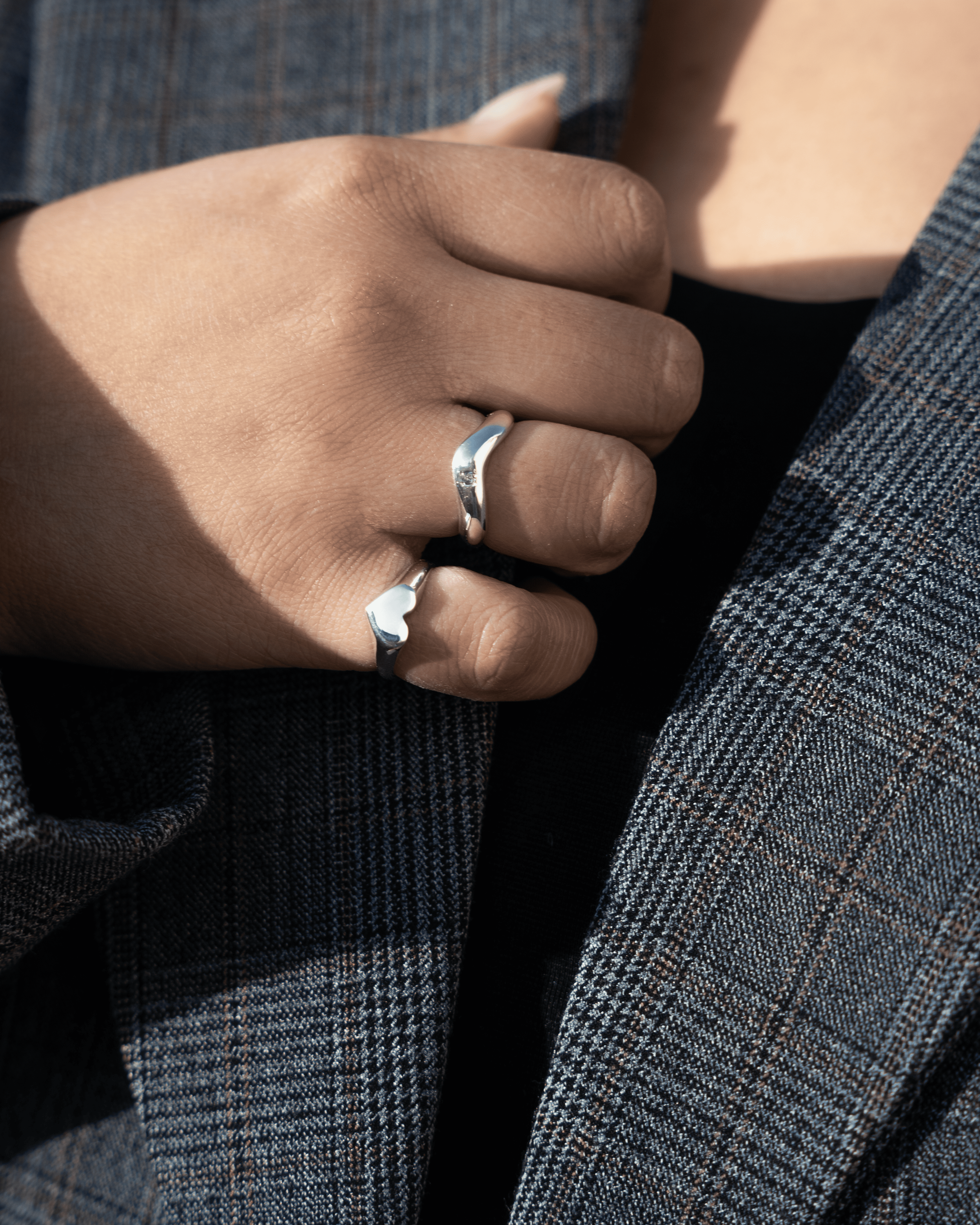 Close-up of a person's hand with a silver ring on the middle finger, resting on their chest with a plaid blazer and black shirt.