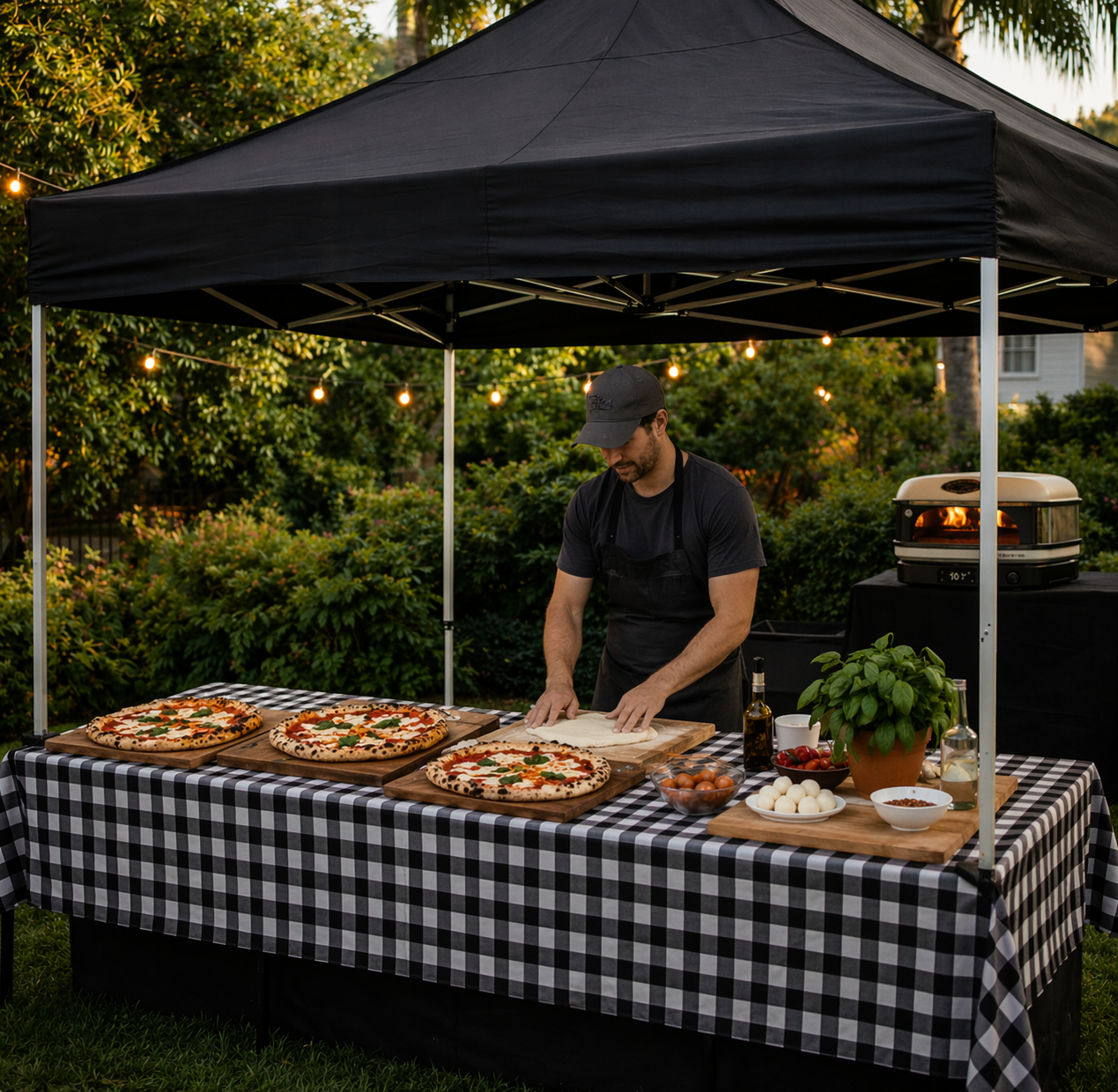 Man making pizza at outdoor pizza station with three finished pizzas on cutting boards, surrounded by ingredients and cooking equipment under a black canopy