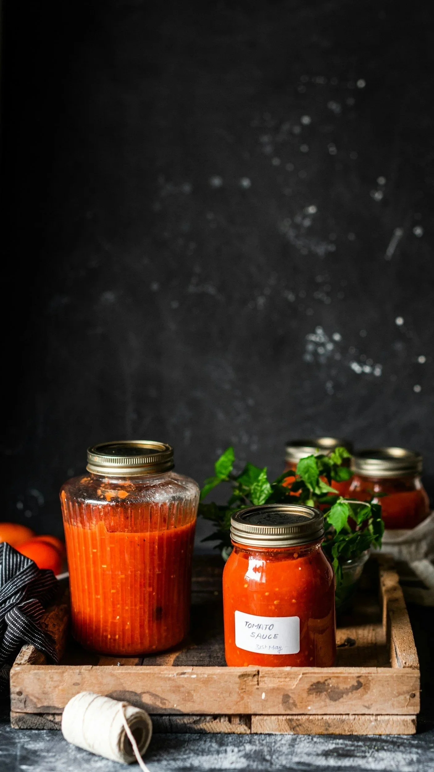 Jars of homemade tomato sauce in a wooden crate with fresh basil and tomatoes, on a dark background.