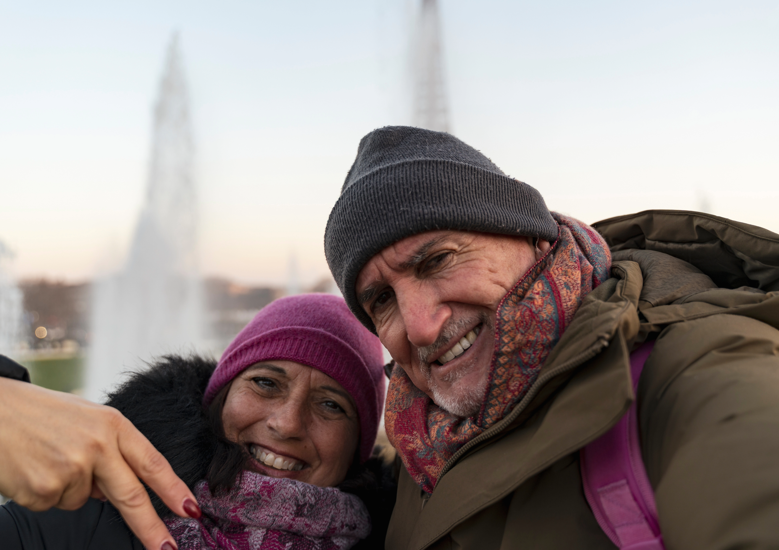 Smiling man and woman wearing winter clothing taking a selfie outdoors with a fountain in the background.