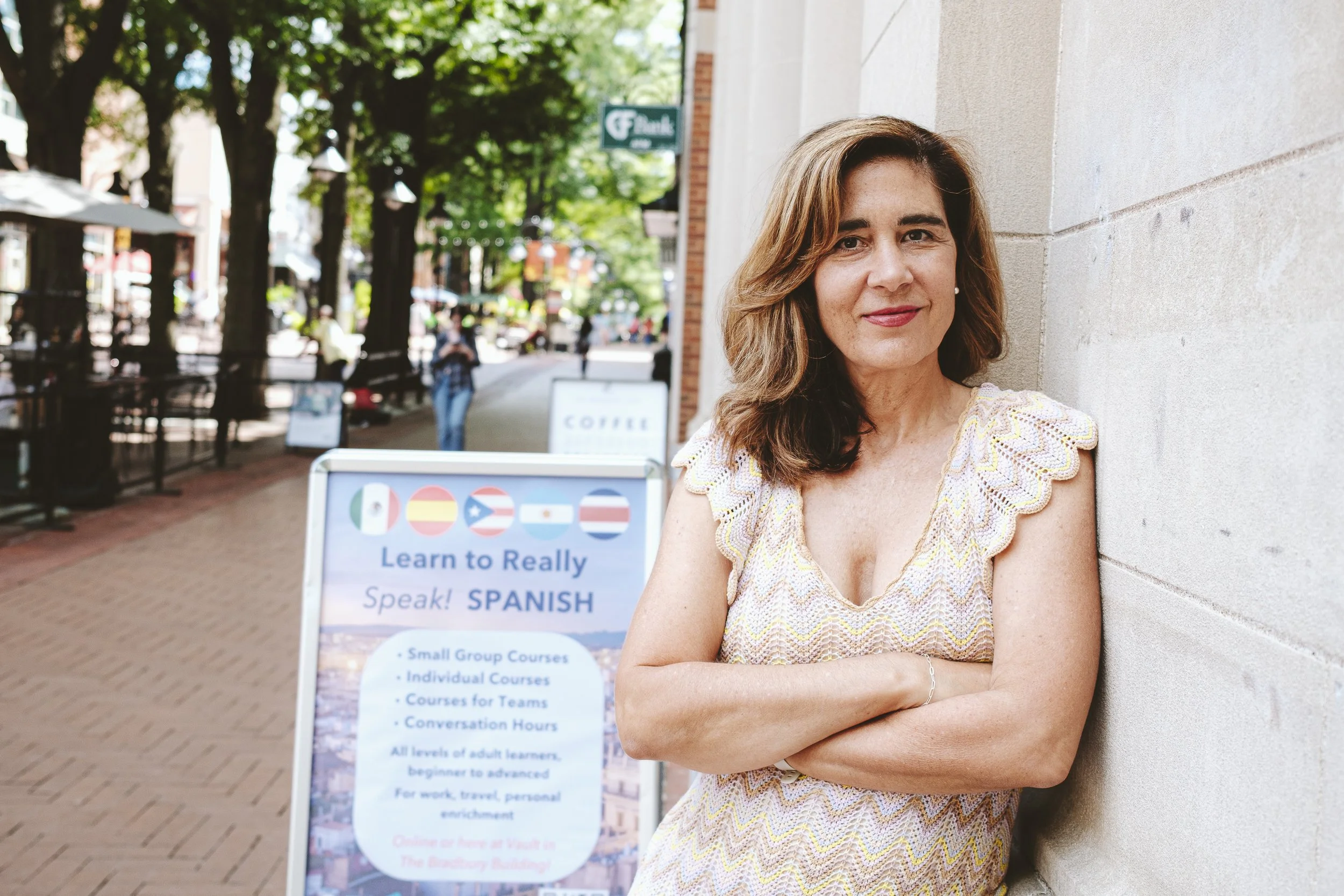 A woman with brown hair, wearing a sleeveless yellow and white dress, leaning against a stone wall on a city sidewalk, with a sign advertising Spanish language courses in the background.