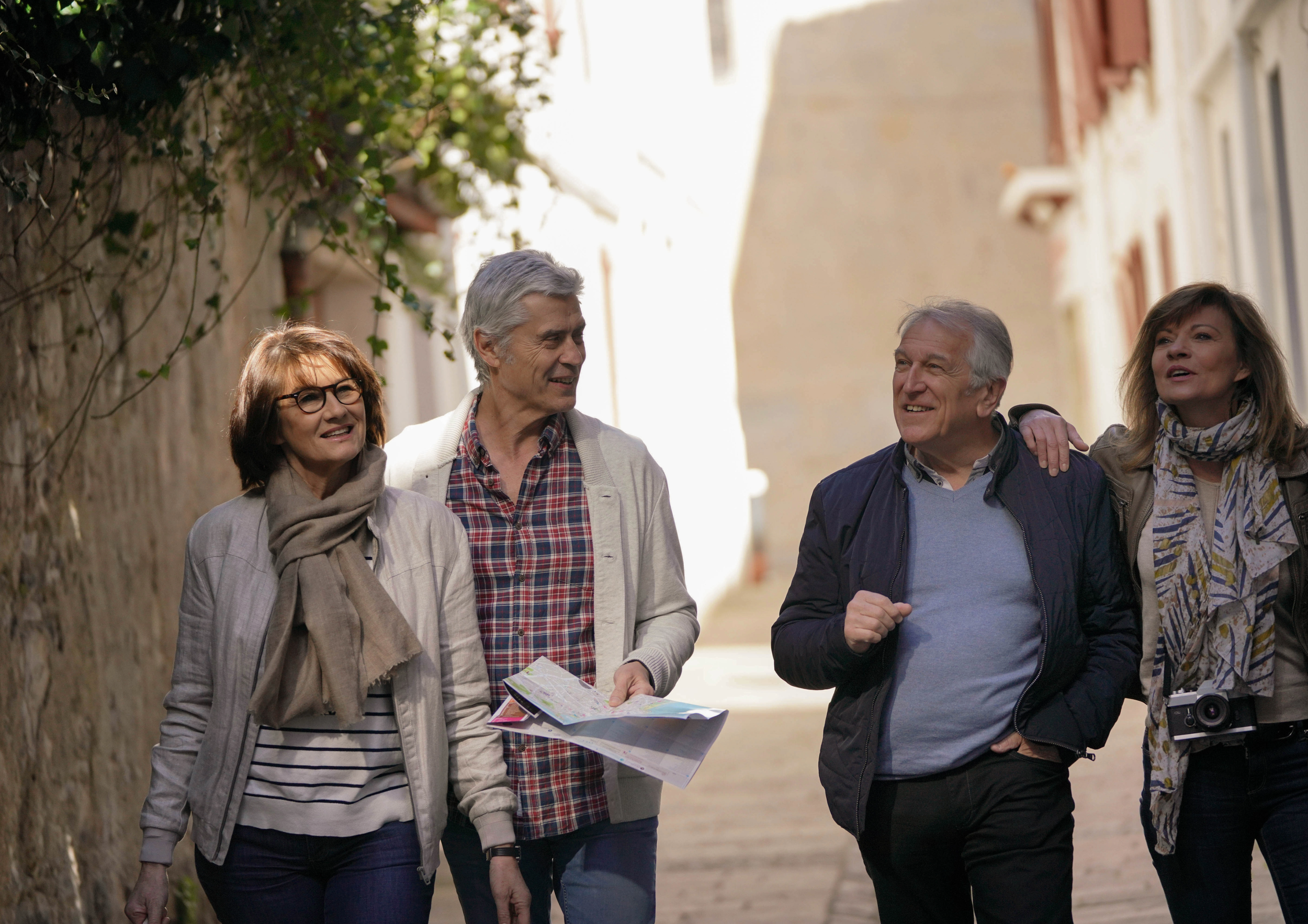 Four mature friends walking together on a sunny street, talking and smiling, one woman holding a map, another woman with a camera around her neck.