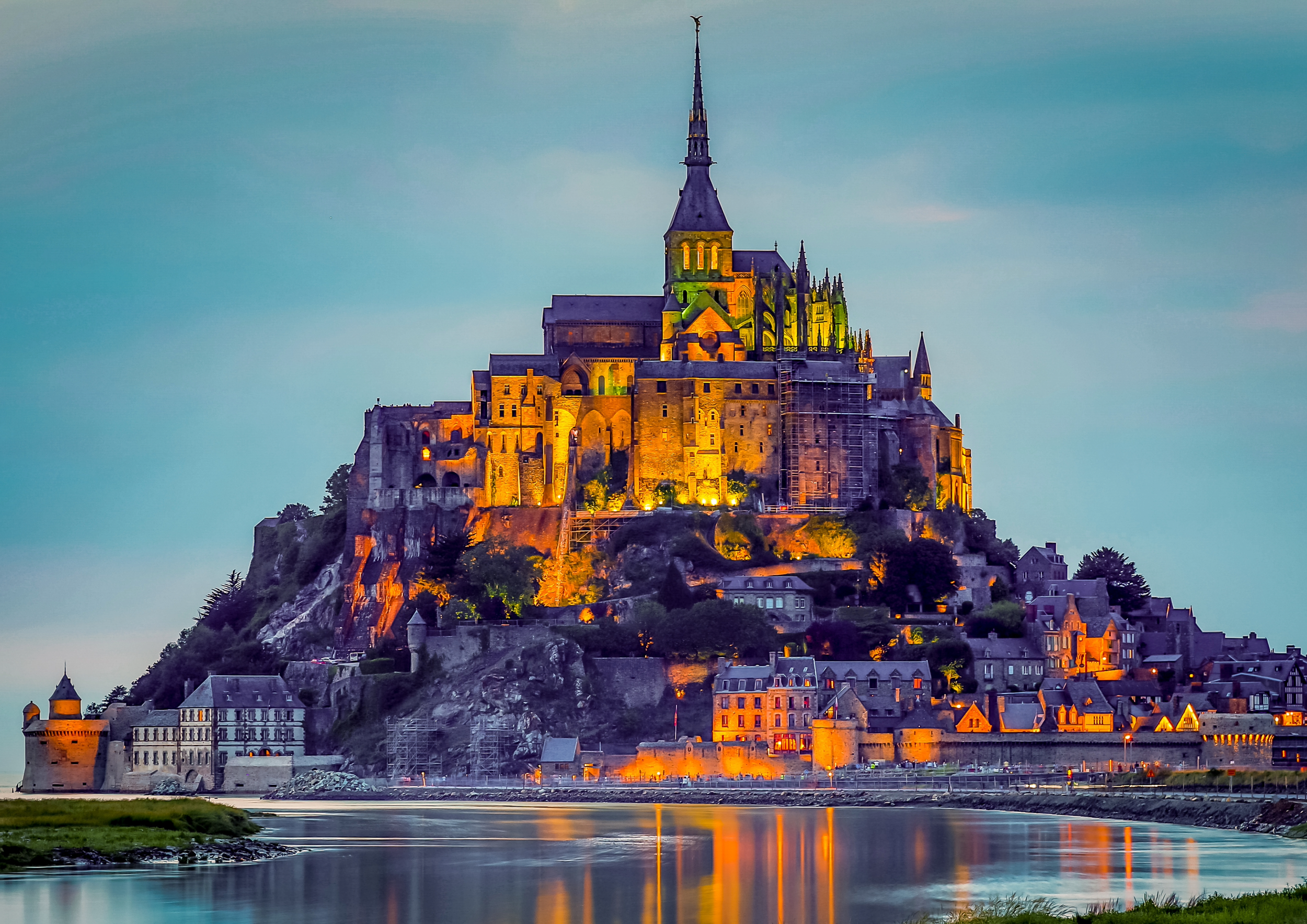 Night view of Mont Saint-Michel, a historic island abbey in France, illuminated with lights, reflected in water.