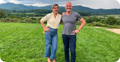 A smiling couple standing on a grassy field with mountains in the background.