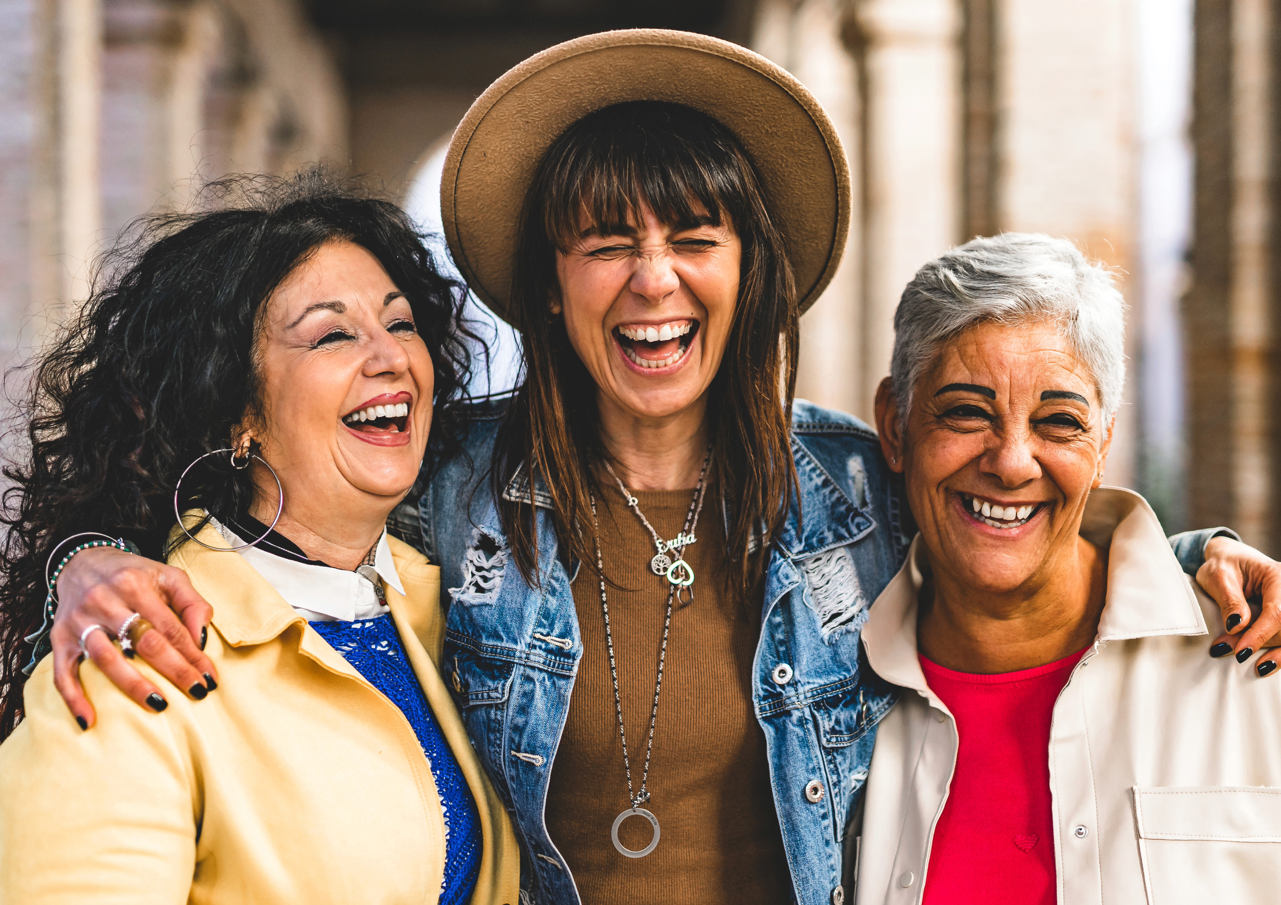 Three women laughing and hugging each other outdoors under a brick archway.
