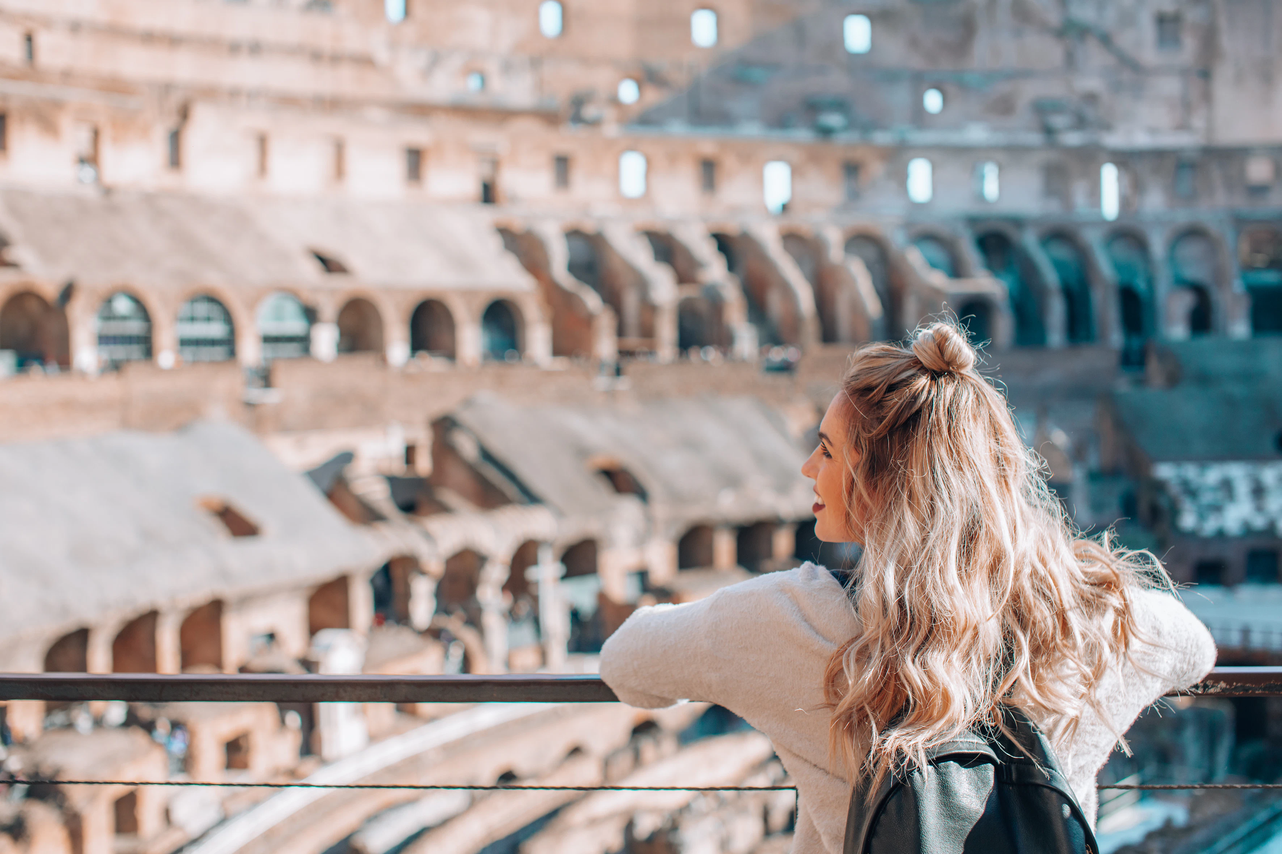 A young woman with long wavy blonde hair, wearing a white sweater and black backpack, smiling and looking at the ancient Colosseum in Rome, Italy.