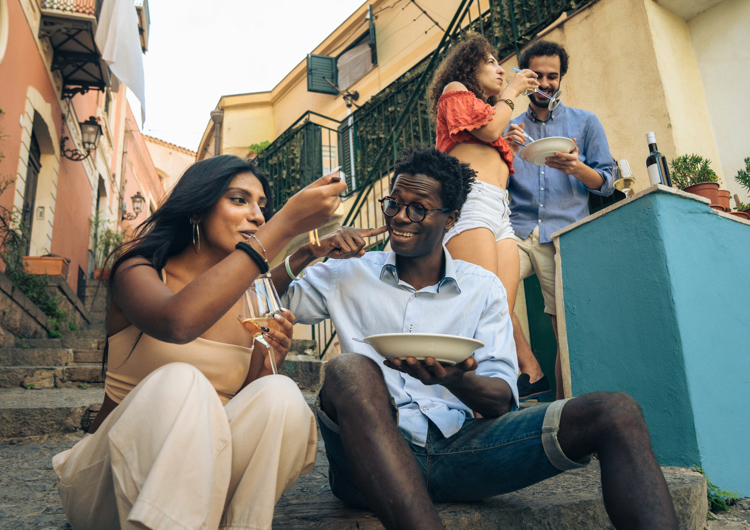 A group of five friends enjoying a gathering on a colorful outdoor staircase in a lively neighborhood, some eating, drinking, and talking.