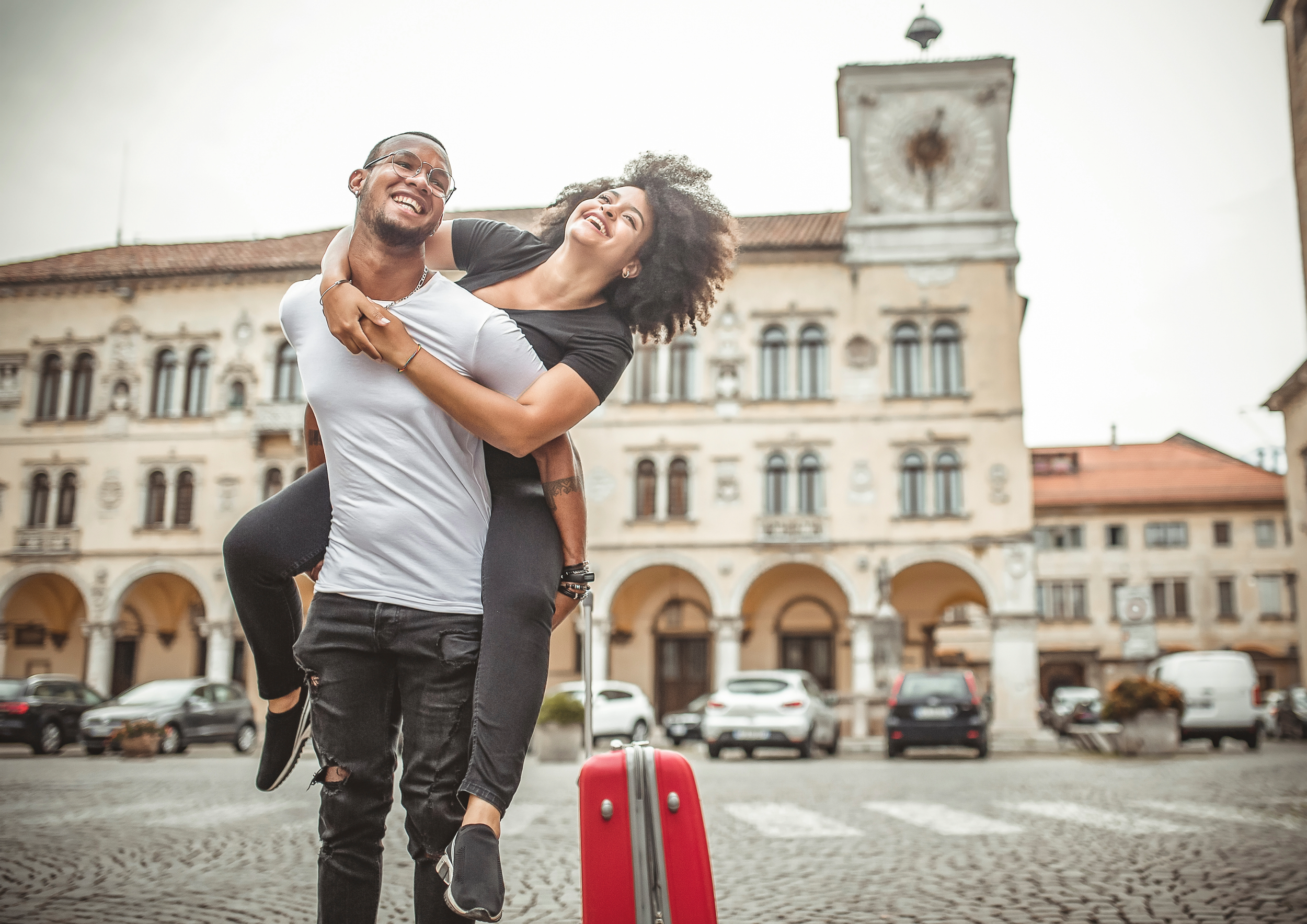 A happy young couple enjoying a piggyback ride on a cobblestone street in front of a historic European building with arches, windows, and a clock tower, with cars parked along the street and a red suitcase in the foreground.