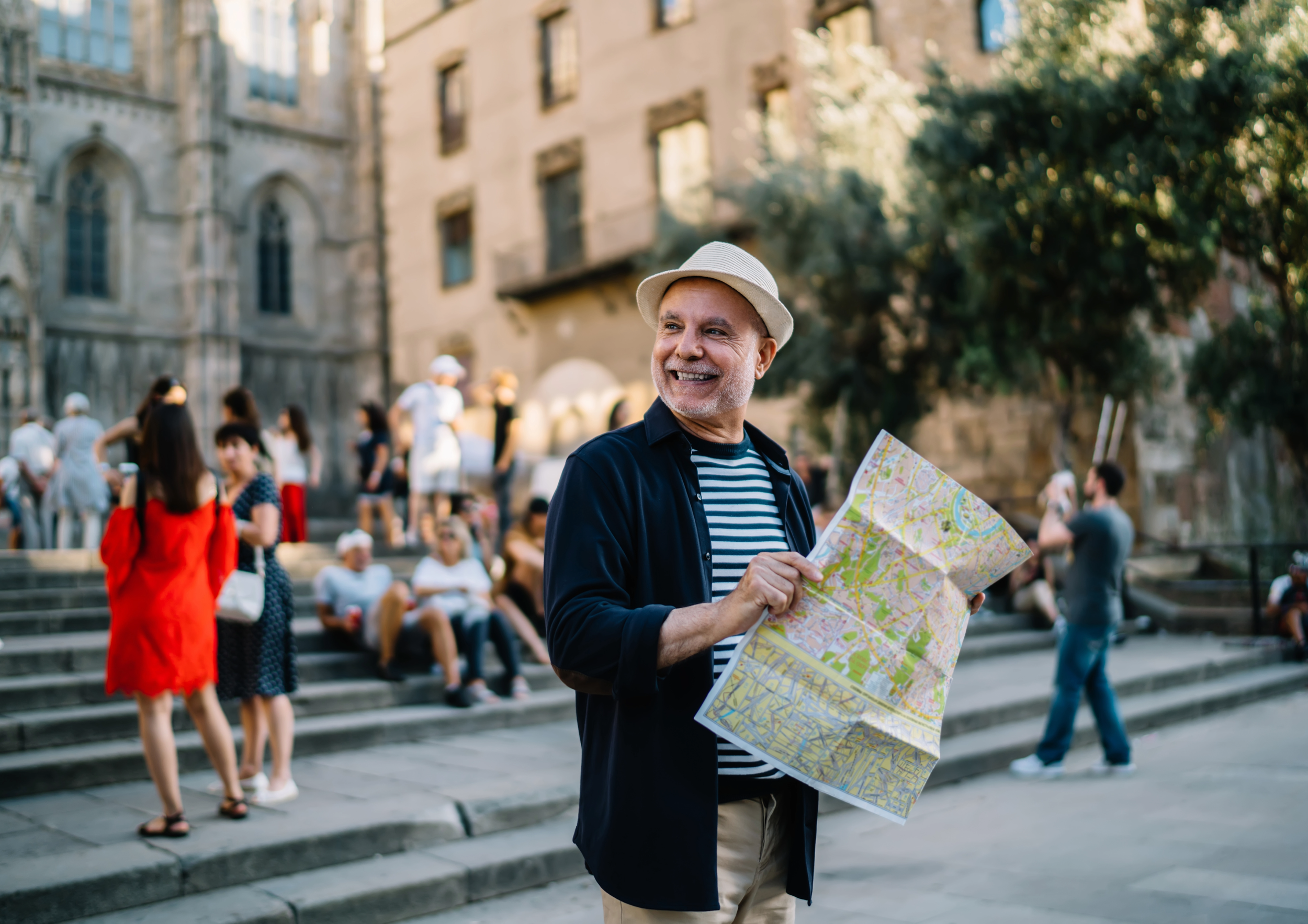 A smiling man with a fedora holding a city map, standing on steps outside a historic building with tourists sitting and walking around in a busy urban area.