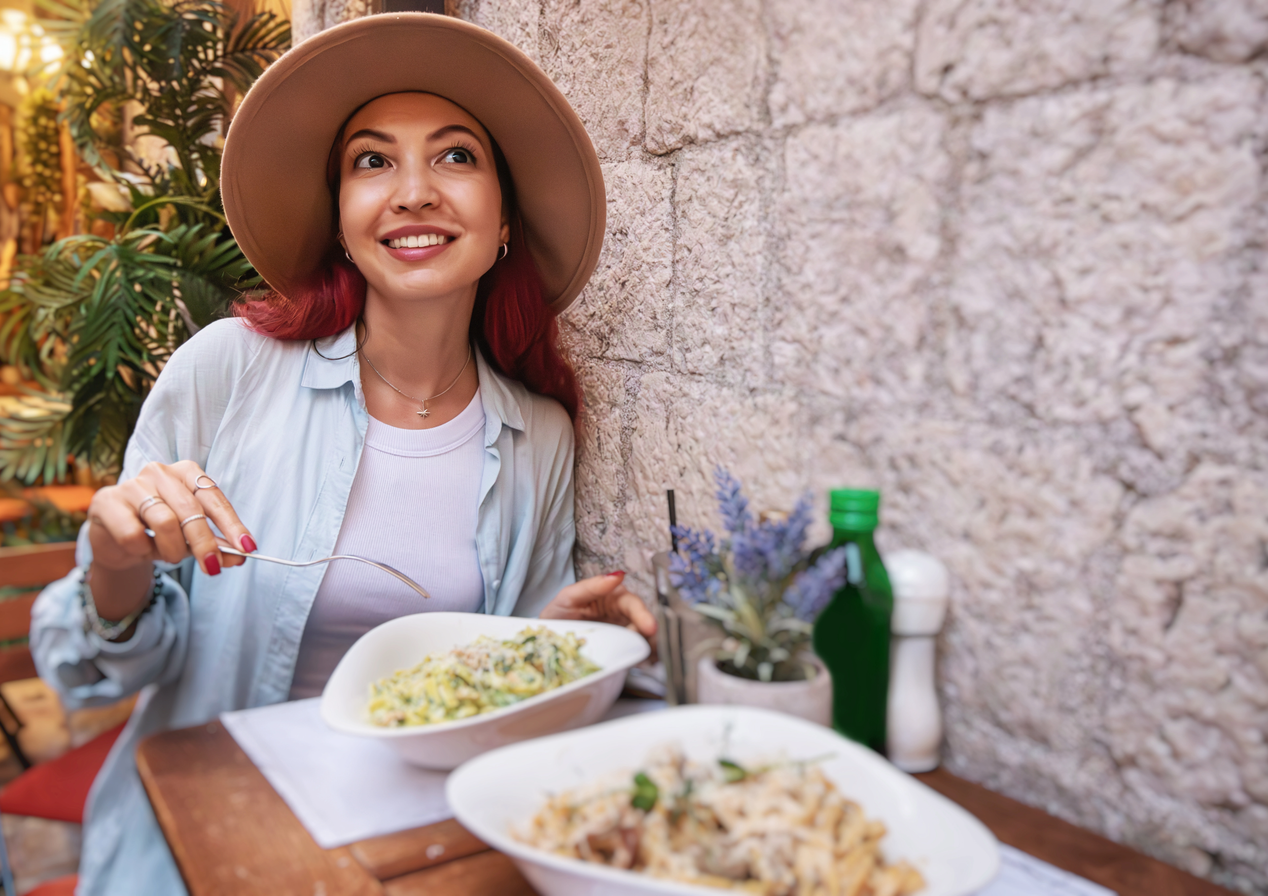 A woman with red hair and a wide-brimmed hat smiling while sitting at an outdoor table with bowls of pasta, a small plant with purple flowers, and condiment bottles, next to a stone wall.