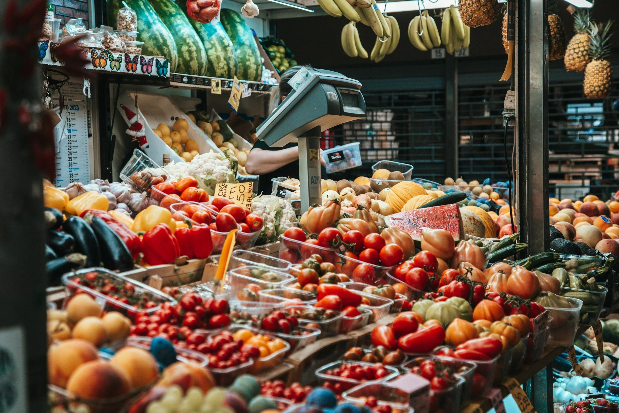 A vendor's stall at a produce market displaying various fresh fruits and vegetables, including tomatoes, peppers, zucchini, melons, pineapples, and other produce, with a person behind the stall checking an electronic scale.