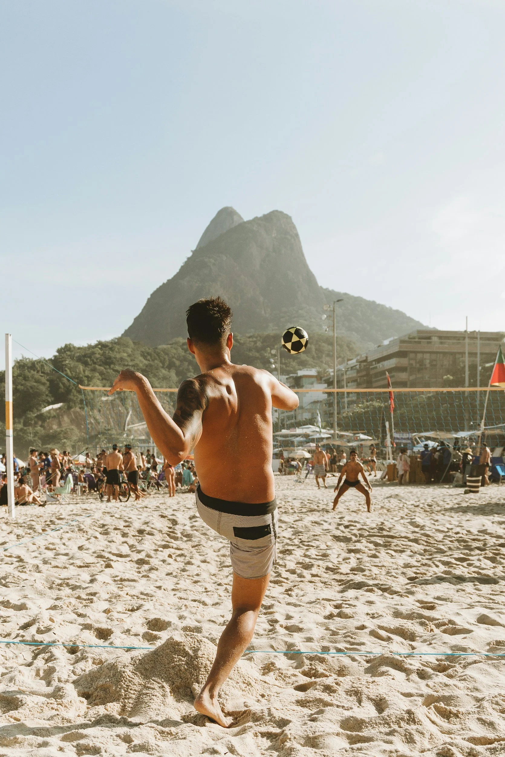 A man playing beach volleyball on a crowded sandy beach with a mountain and buildings in the background.