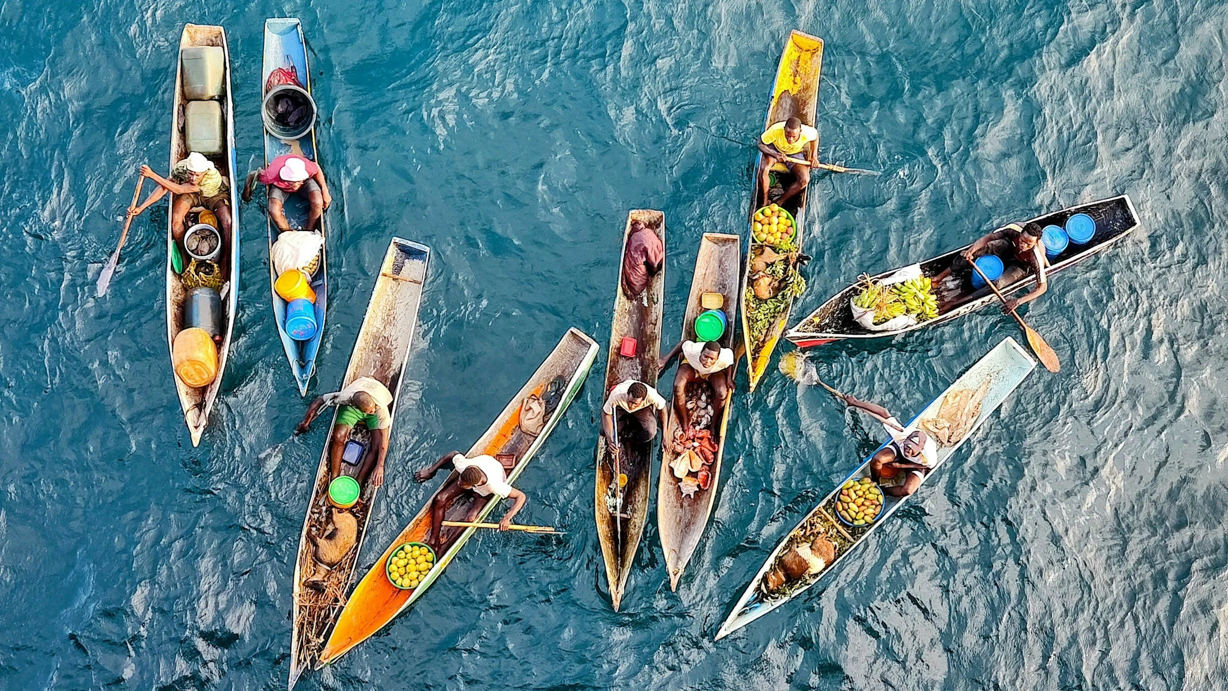Aerial view of multiple colorful fishing boats with people on the water, some rowing and some sitting with fishing equipment and catches visible.
