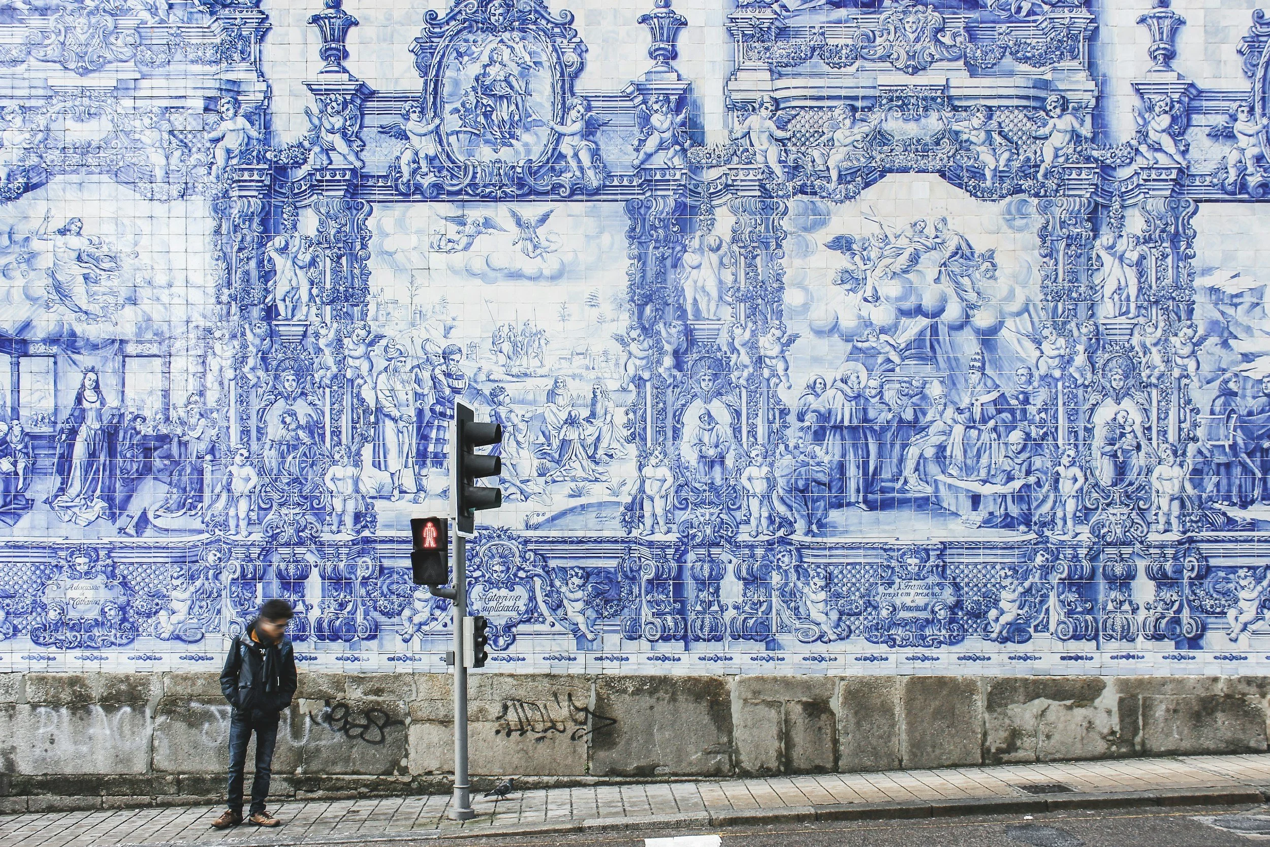 A man in a black jacket and face mask stands on a sidewalk beneath a traffic light with a red pedestrian signal. Behind him is a large blue and white tile mural depicting various historical and religious scenes. The wall is weathered at the bottom with some graffiti.