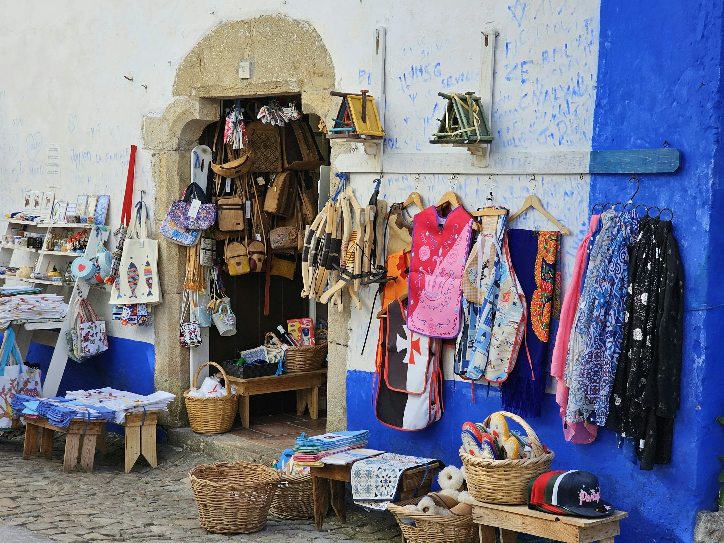 Street market stall displaying colorful bags, aprons, clothes, and accessories against a blue-painted wall.
