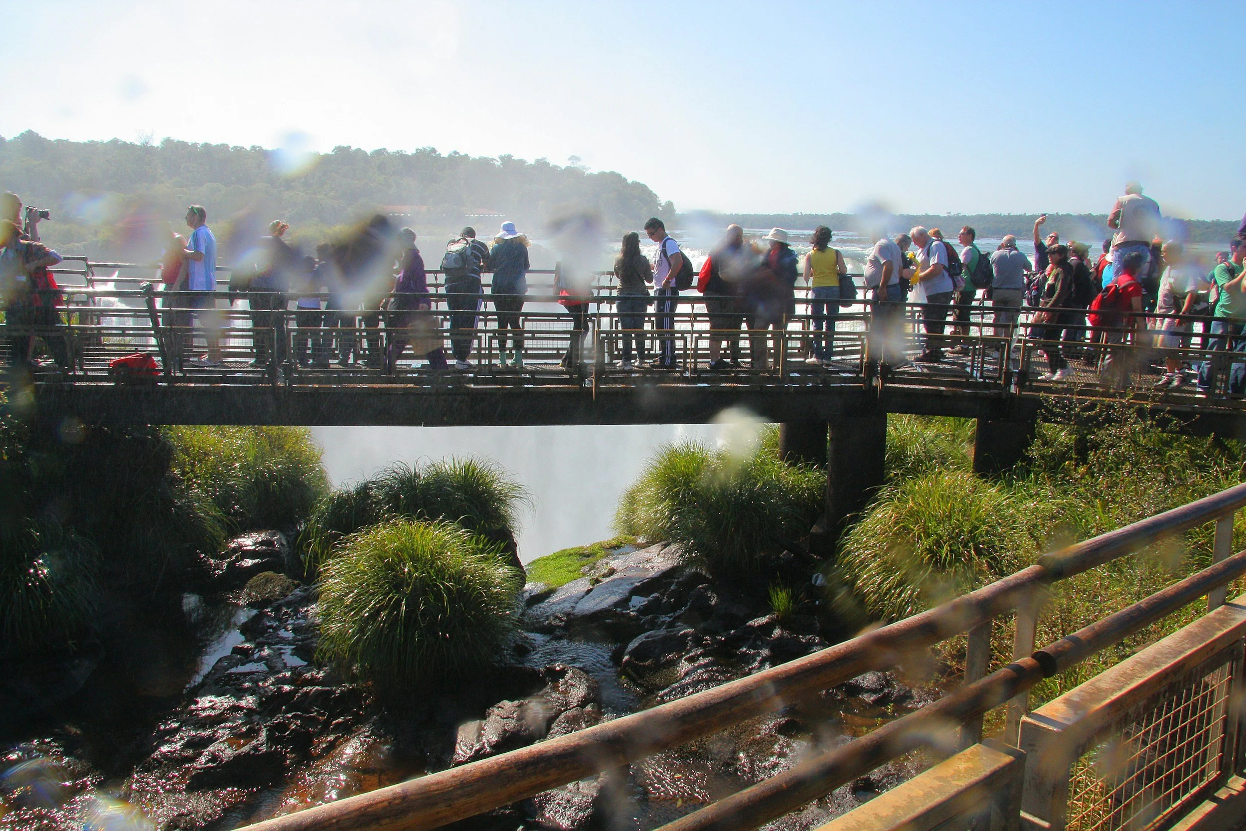 Group of people standing on a bridge overlooking a waterfall with mist and greenery in the background.