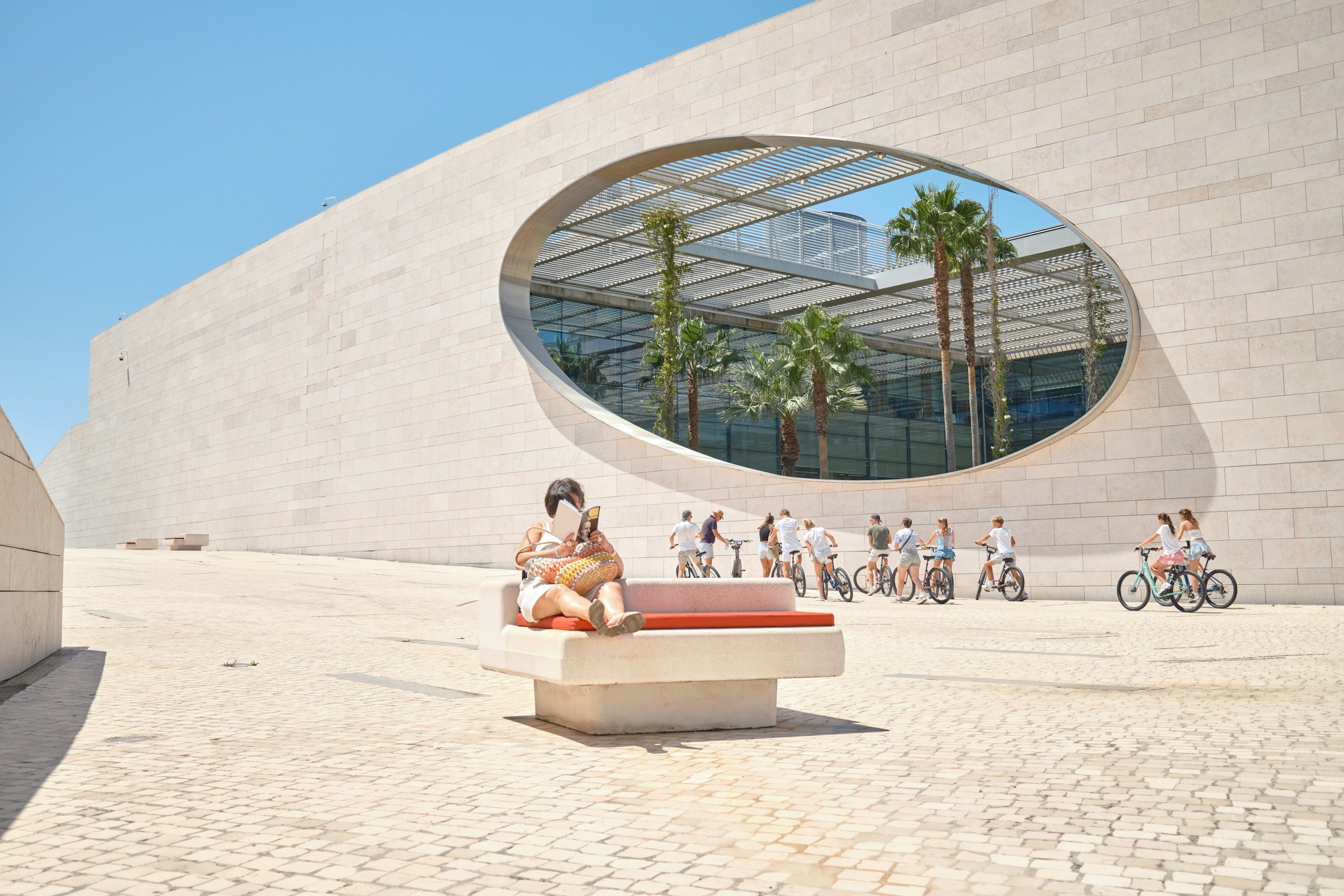 A modern building with an oval window and palm trees inside. A woman sitting on a bench reading a book, with a group of people riding bicycles in the background on a sunny day.