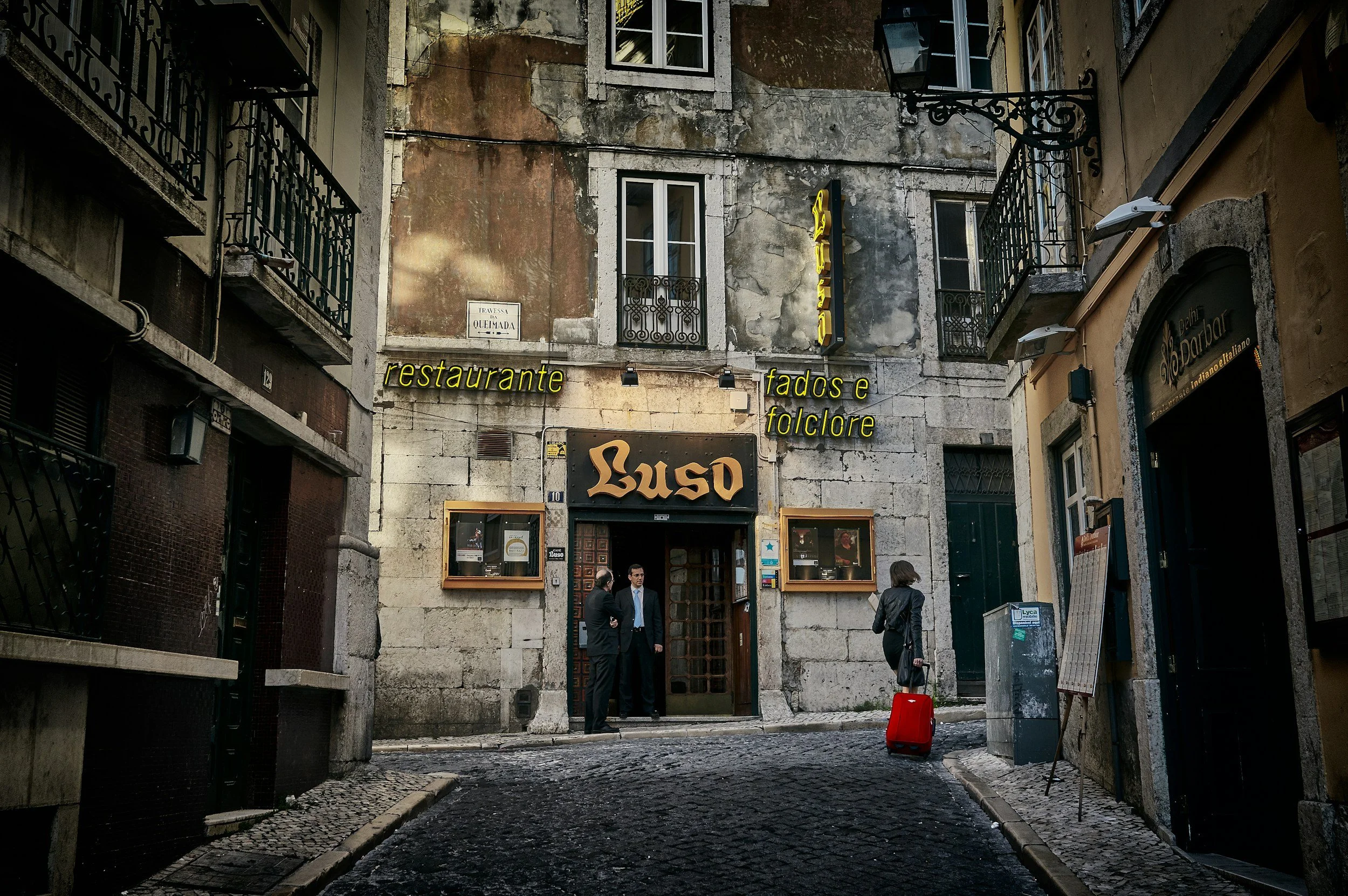 Front of a rustic restaurant named Luso with a neon sign, located on a narrow cobblestone street. Two men in suits stand near the entrance, and a woman with a red suitcase stands nearby, all in an old European neighborhood.