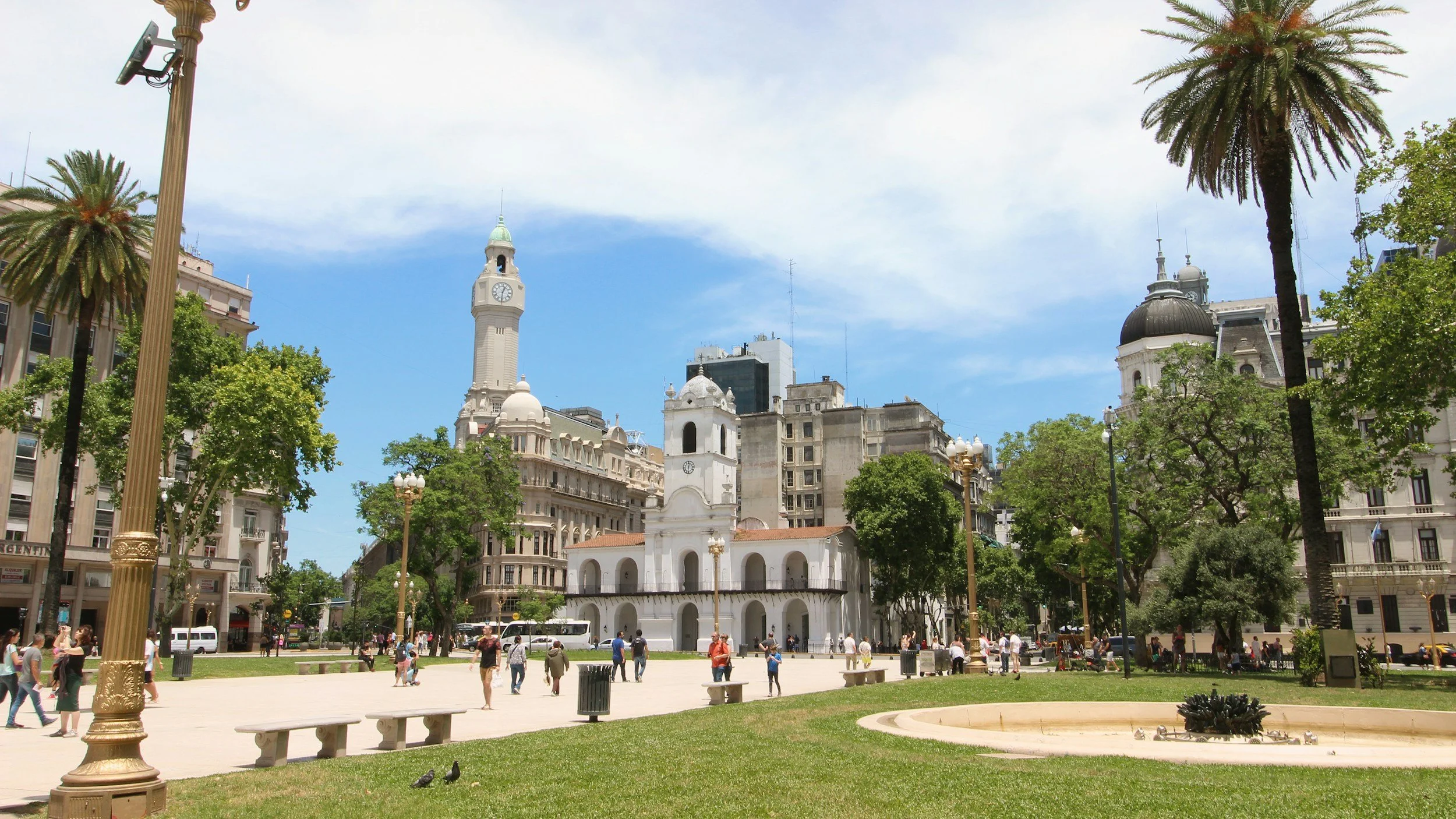 City square in Buenos Aires with historic buildings, a clock tower, people walking, trees, and a park with benches and a fountain.