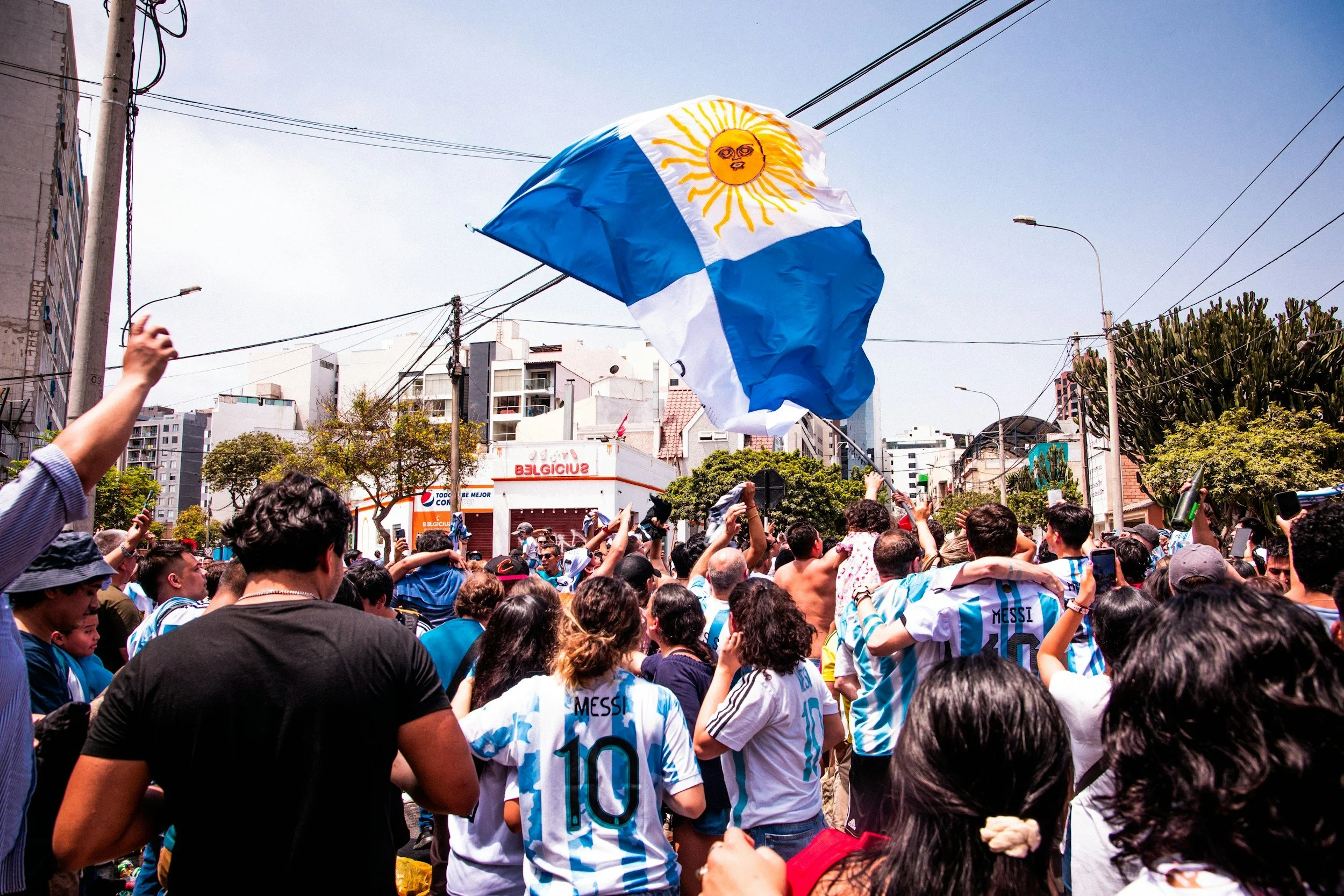 Crowd of people wearing Argentina soccer jerseys in a street, cheering and waving flags, with one person holding a large Argentine flag with a sun emblem, in an urban setting with buildings and power lines.
