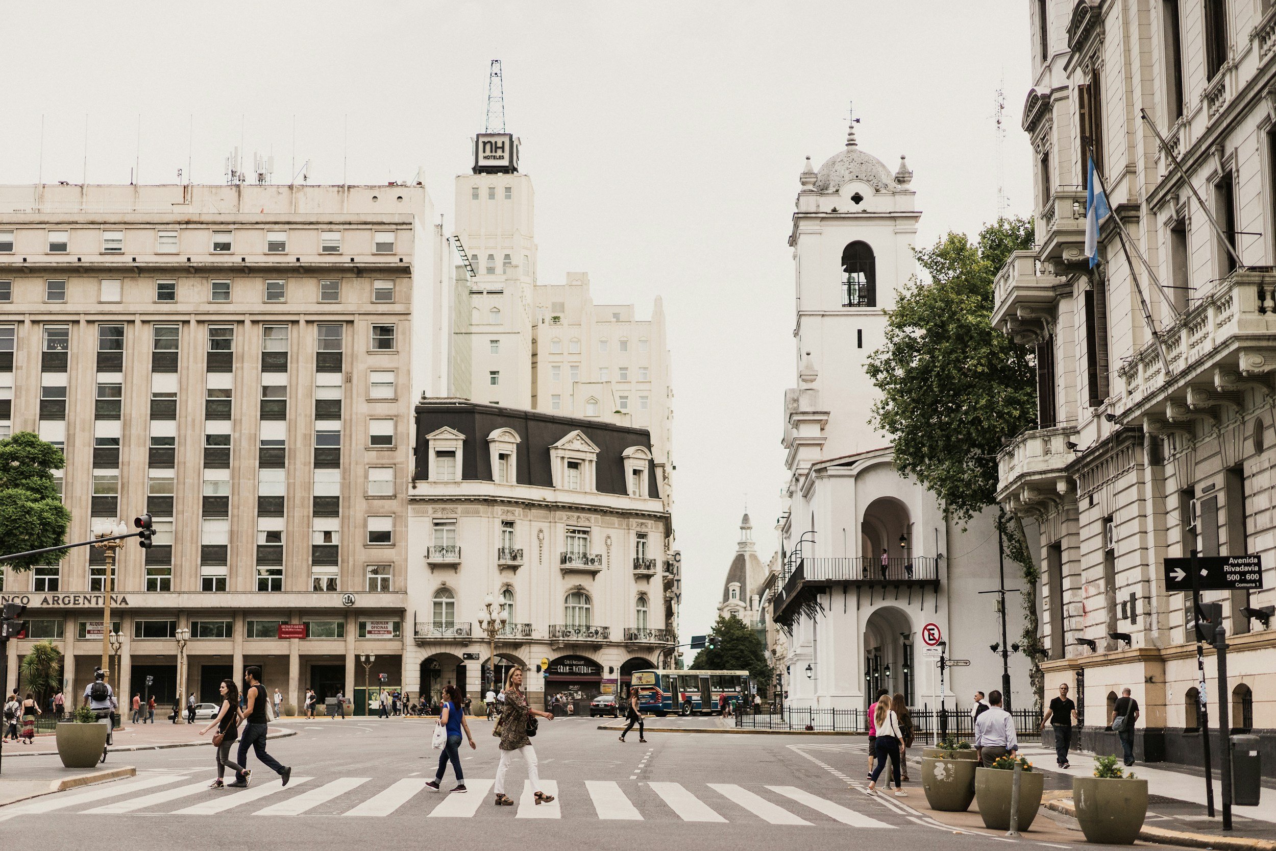 City scene with people crossing a street, white historic buildings with columns, trees, a bus, and a cloudy sky.