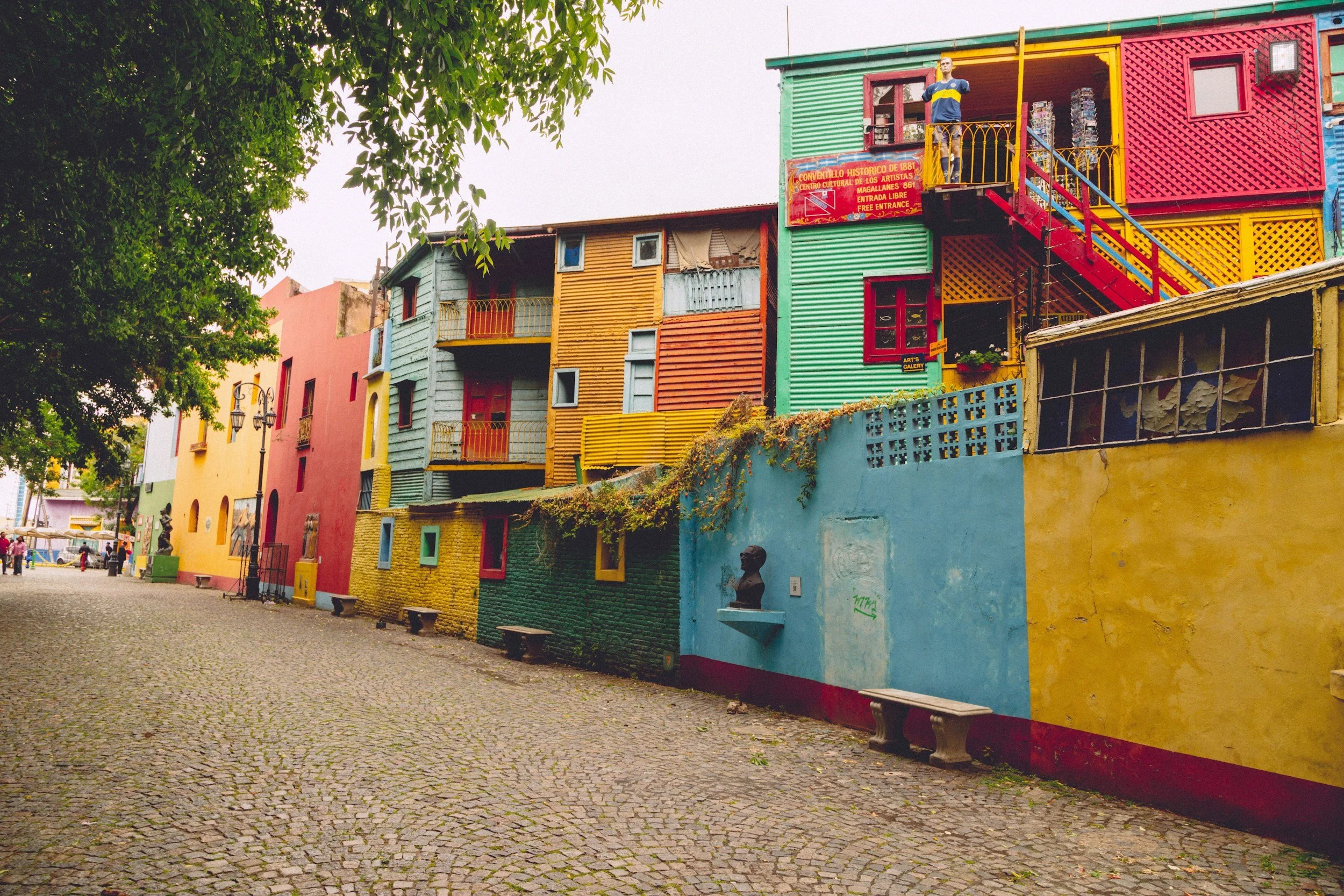Colorful buildings in La Boca neighborhood, Buenos Aires, Argentina, with painted walls, small balconies, and benches along cobblestone street.