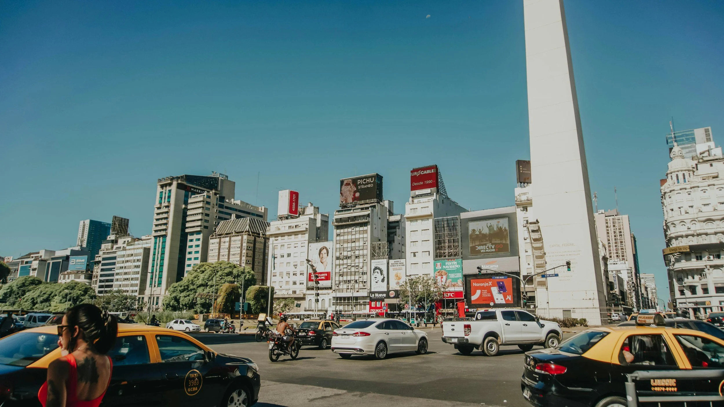 City street with tall buildings, billboards, cars, taxis, motorcycles, and a woman in the foreground, under a clear blue sky.