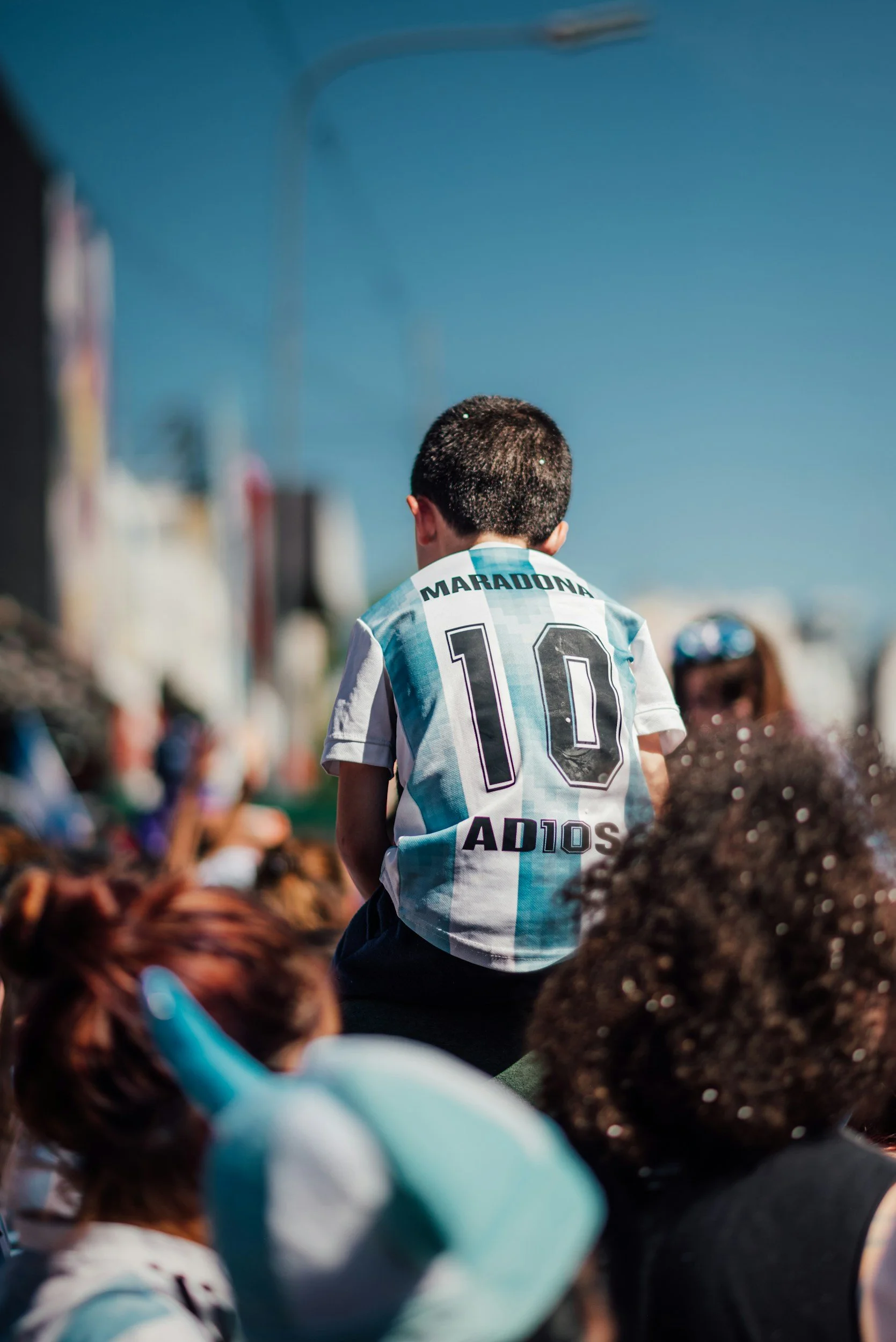 A young boy wearing an Argentina soccer jersey with the name 'Maradona' and number 10 on the back, sitting on someone's shoulders at a crowded outdoor event on a bright sunny day.