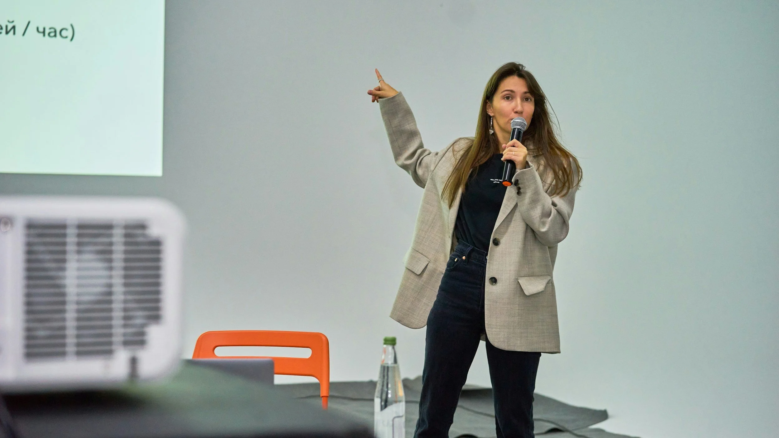 A woman with long brown hair wearing a beige blazer, black shirt, and black pants is speaking into a microphone while pointing at a projected slide in a presentation room. There is an orange chair, a water bottle, and a black table in the background.