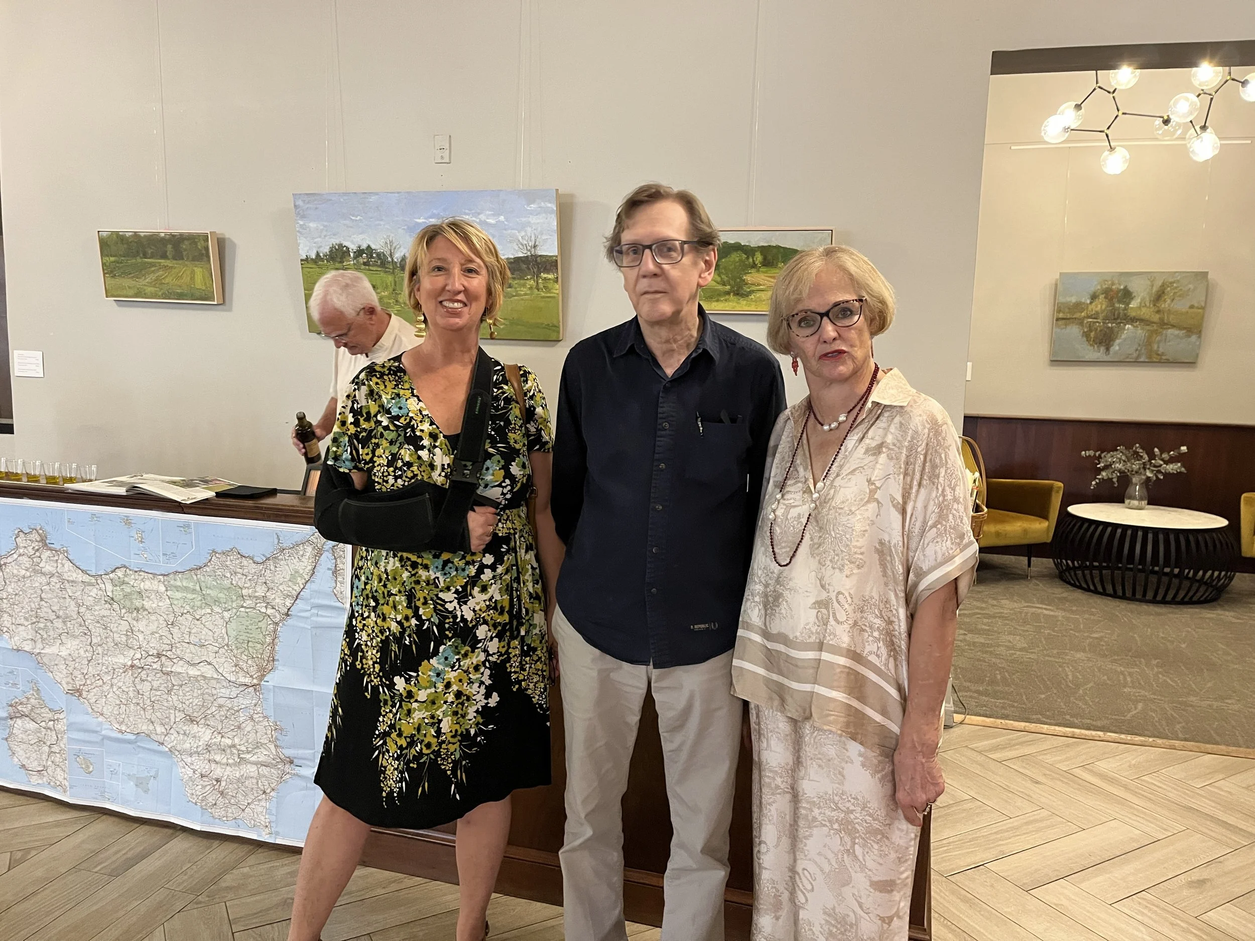 Three people posing in front of landscape paintings.