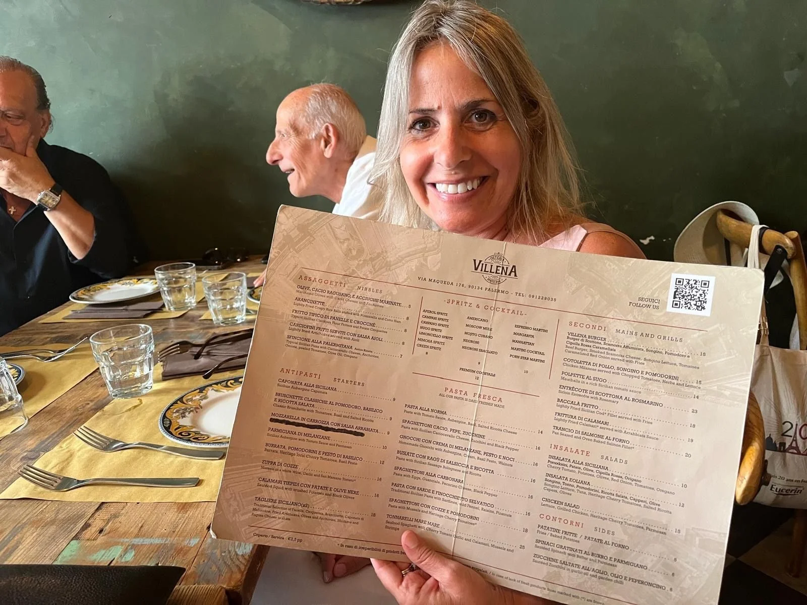 A woman smiling and holding a menu at a dining table in a restaurant, with other diners visible in the background.