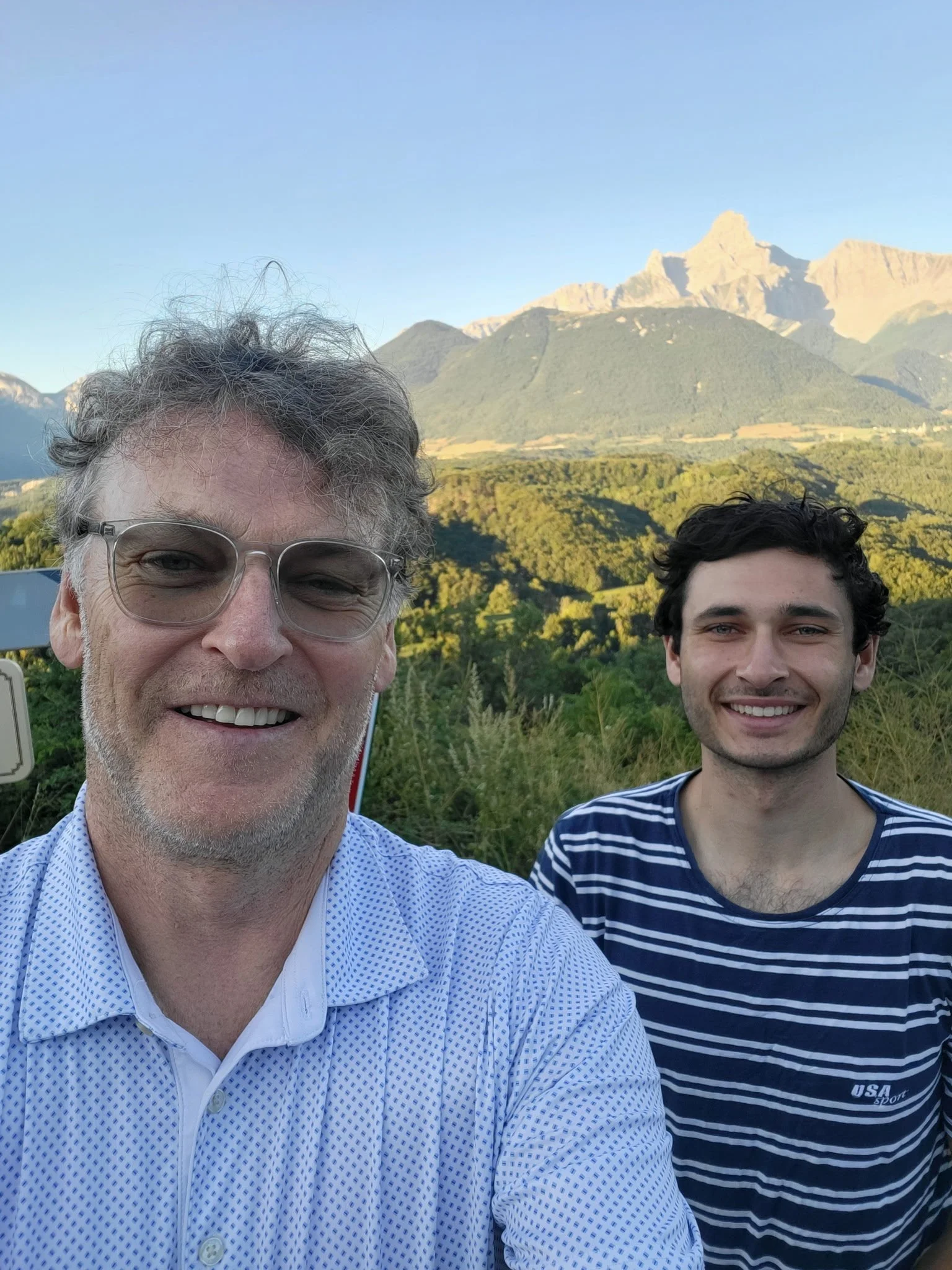 Two men smiling for a selfie outdoors with a mountain and forest in the background. One man has glasses and gray hair, wearing a white patterned shirt; the other has dark curly hair, wearing a striped shirt.