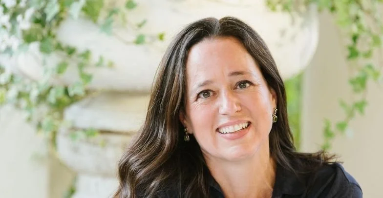 Smiling woman with long dark hair and earrings, outdoors with greenery and a white decorative planter in the background.