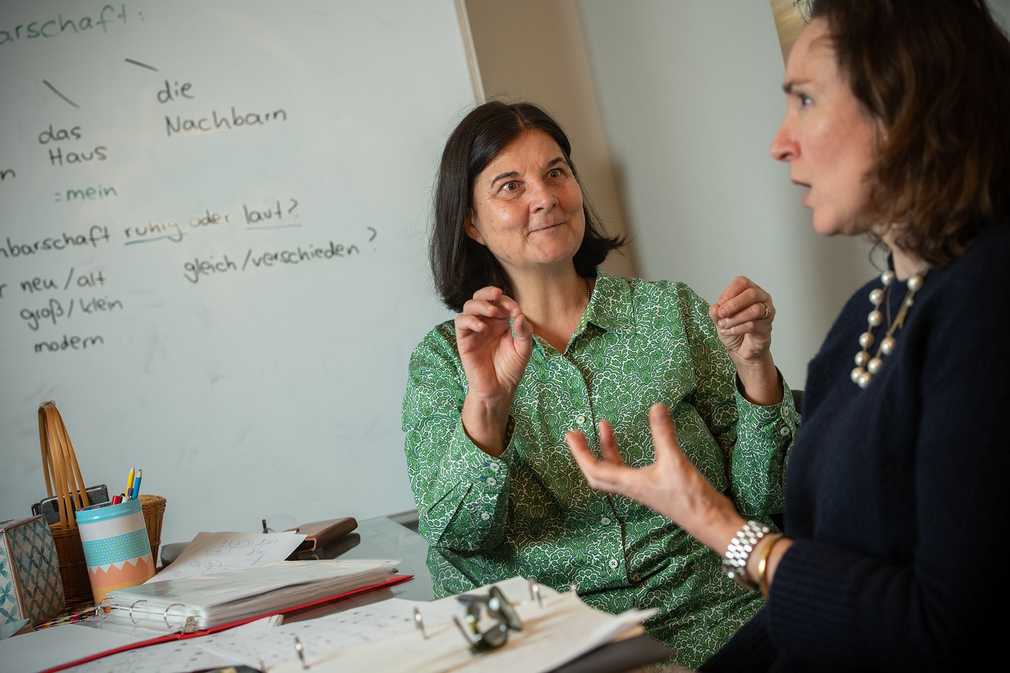 Two women having a discussion in a classroom, with a whiteboard showing German words and phrases in the background.