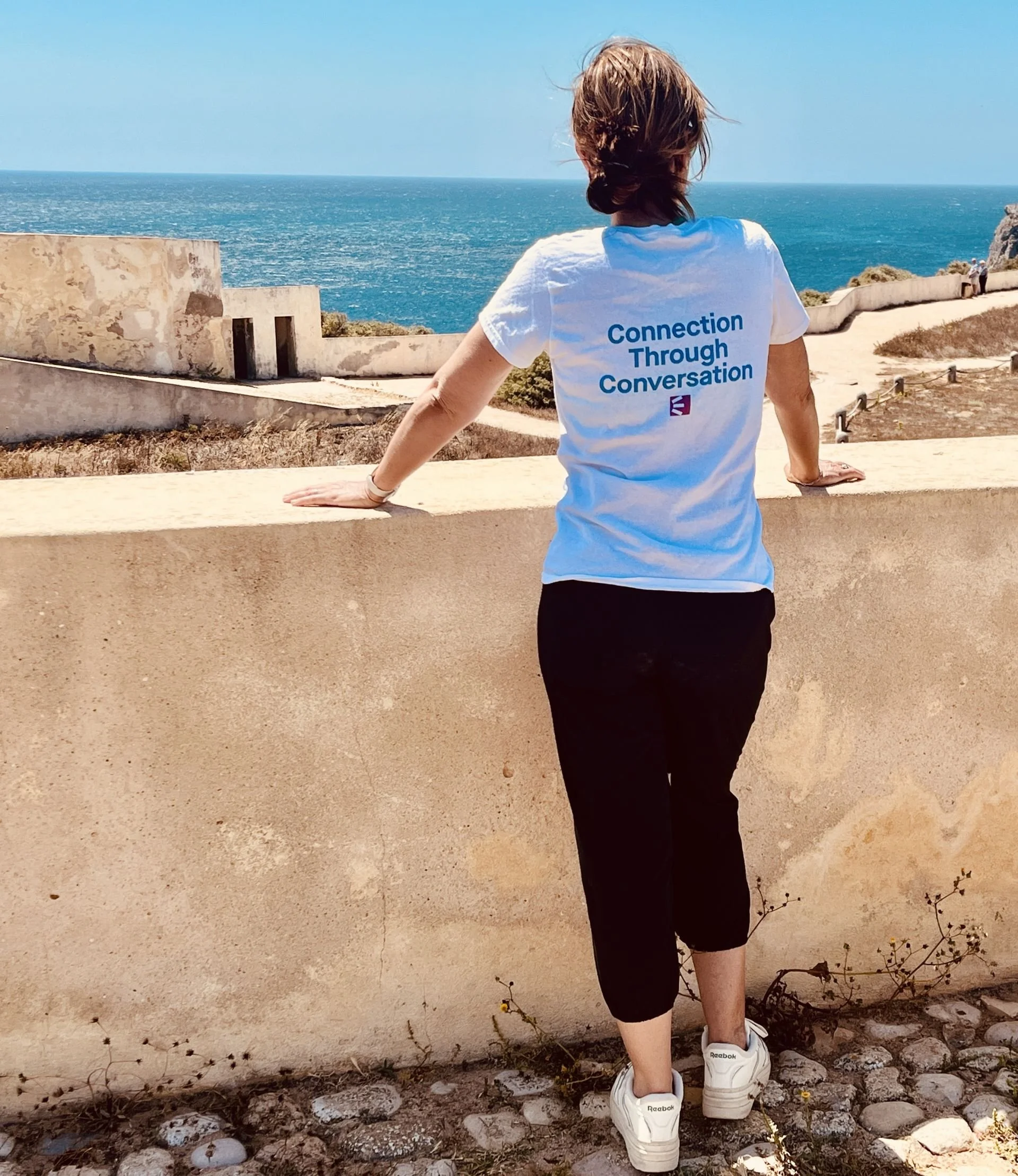 Woman with short hair wearing a white t-shirt with blue text and black pants, standing by a beige wall looking at the ocean.