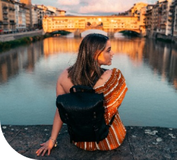 A woman with shoulder-length brown hair sitting by a river at sunset, wearing a striped top and carrying a black backpack, with buildings and a bridge in the background.