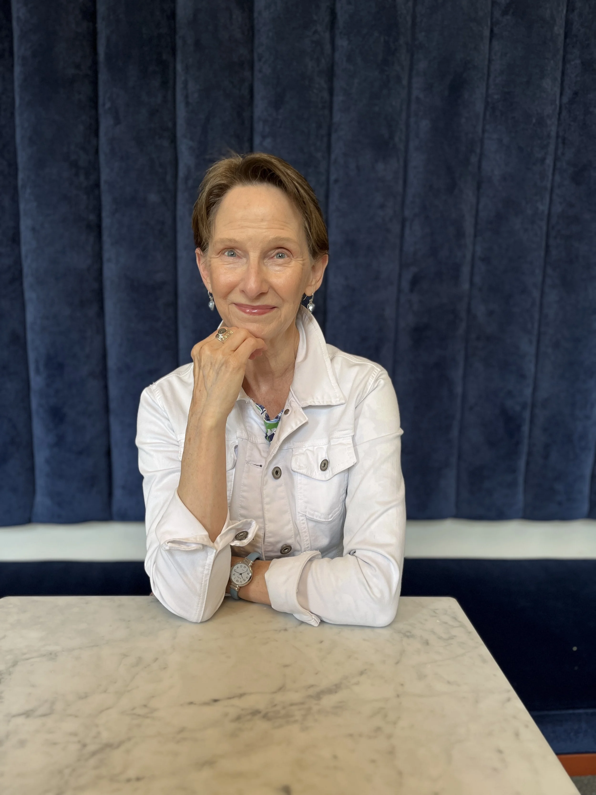A woman with short brown hair sitting at a marble table in front of a velvet blue curtain. She is wearing a white jacket, earrings, and a watch, and is resting her chin on her hand while smiling.