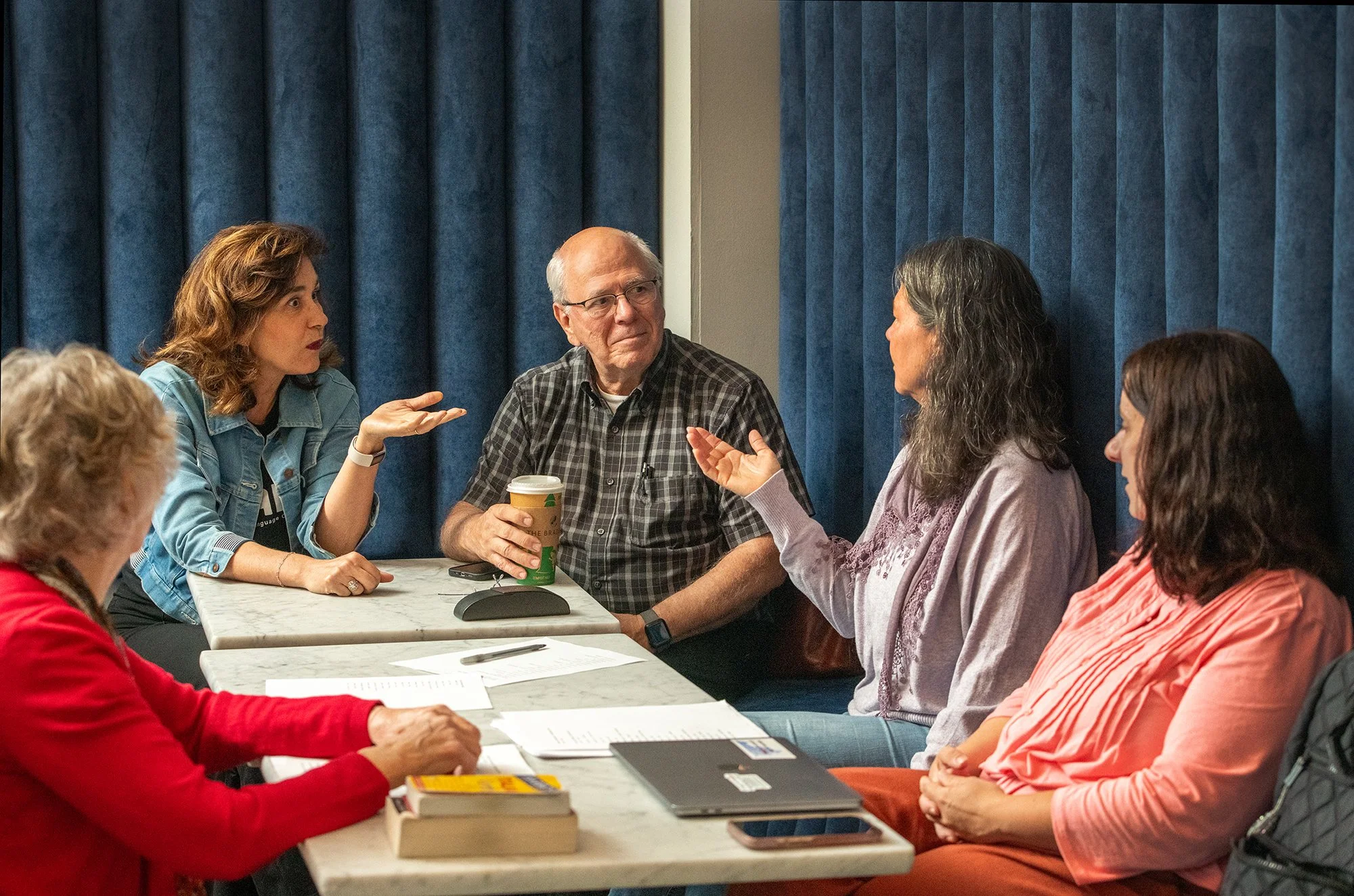 A group of five people sitting around a table in a meeting room, engaging in a discussion. The room has blue curtains in the background, and there are papers, books, a laptop, and a coffee cup on the table.