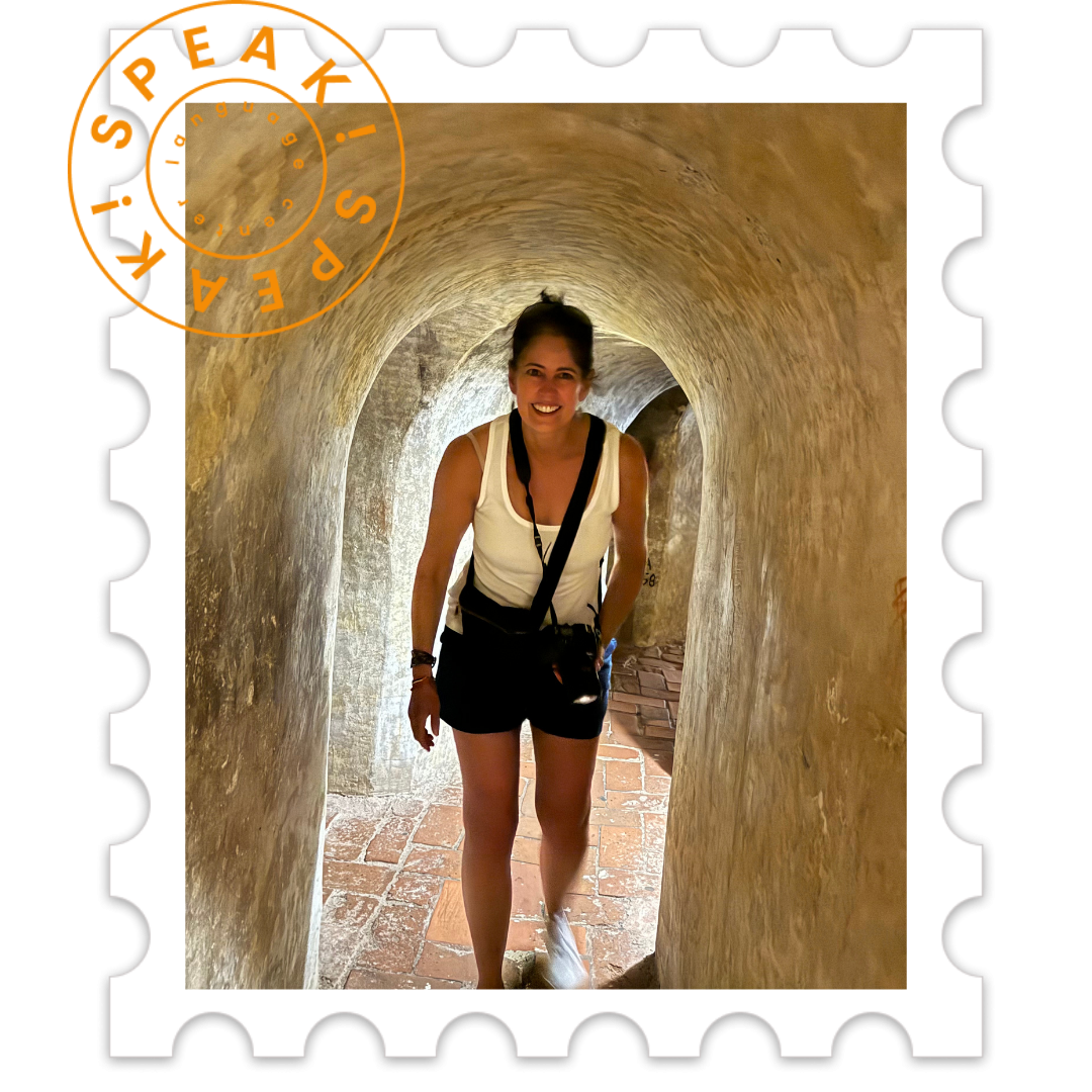 A woman smiling and standing in a narrow, arched brick tunnel with stone walls, holding a camera with a strap around her neck, wearing a white tank top and black shorts.